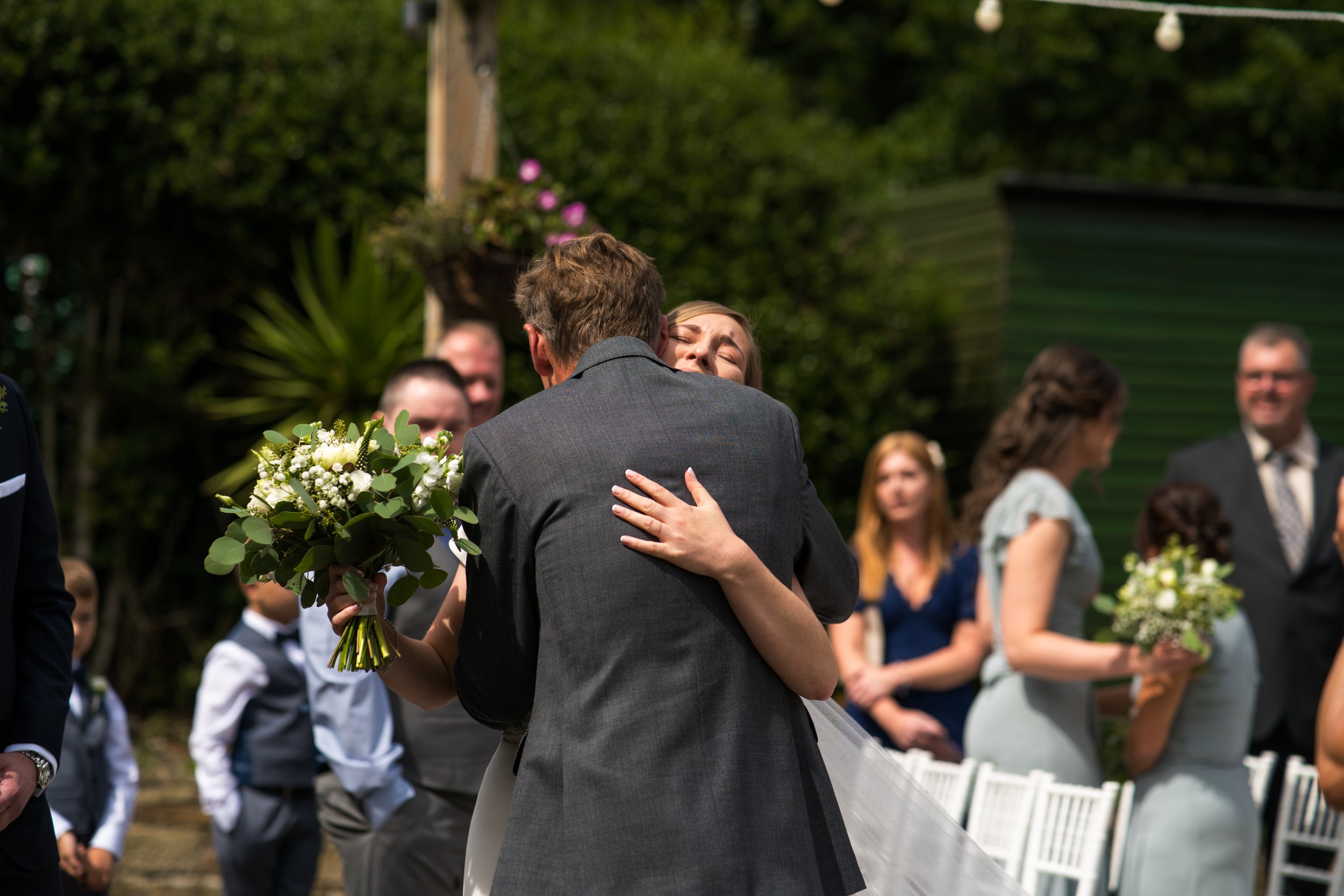 Bride and groom sharing a hug at an outdoor wedding ceremony, with friends and family watching in the background.