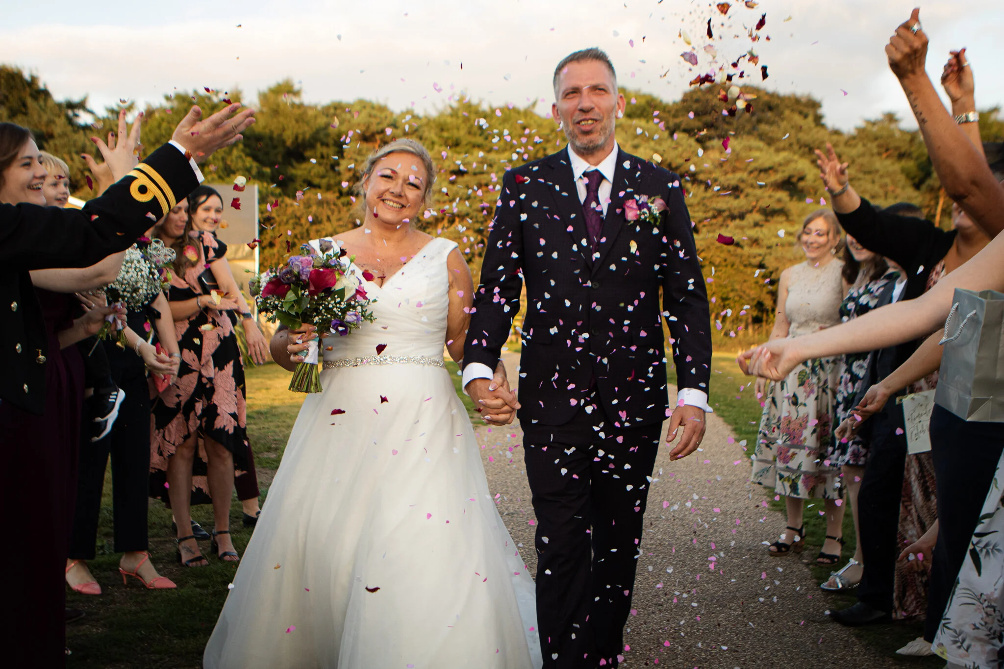 Bride and groom holding hands walking through confetti at outdoor wedding reception with guests celebrating on both sides.