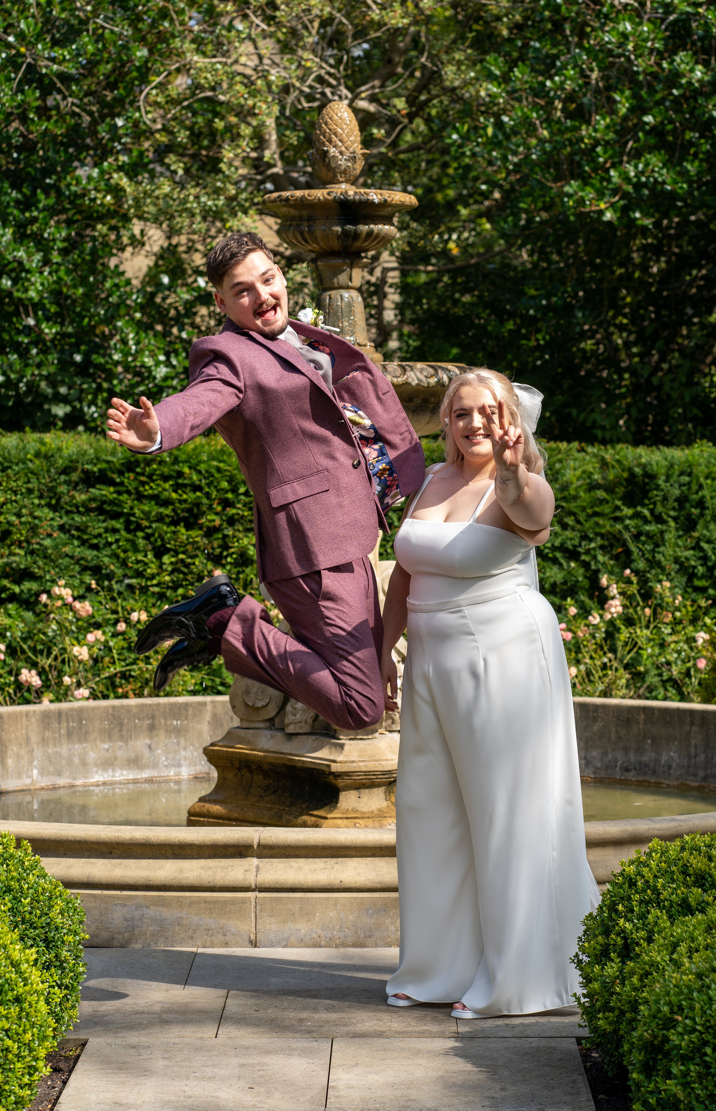 A happy couple in wedding attire posing in front of a fountain in a garden. The groom, dressed in a maroon suit, is jumping with joy, while the bride, in a white wedding dress, is standing and making a peace sign.