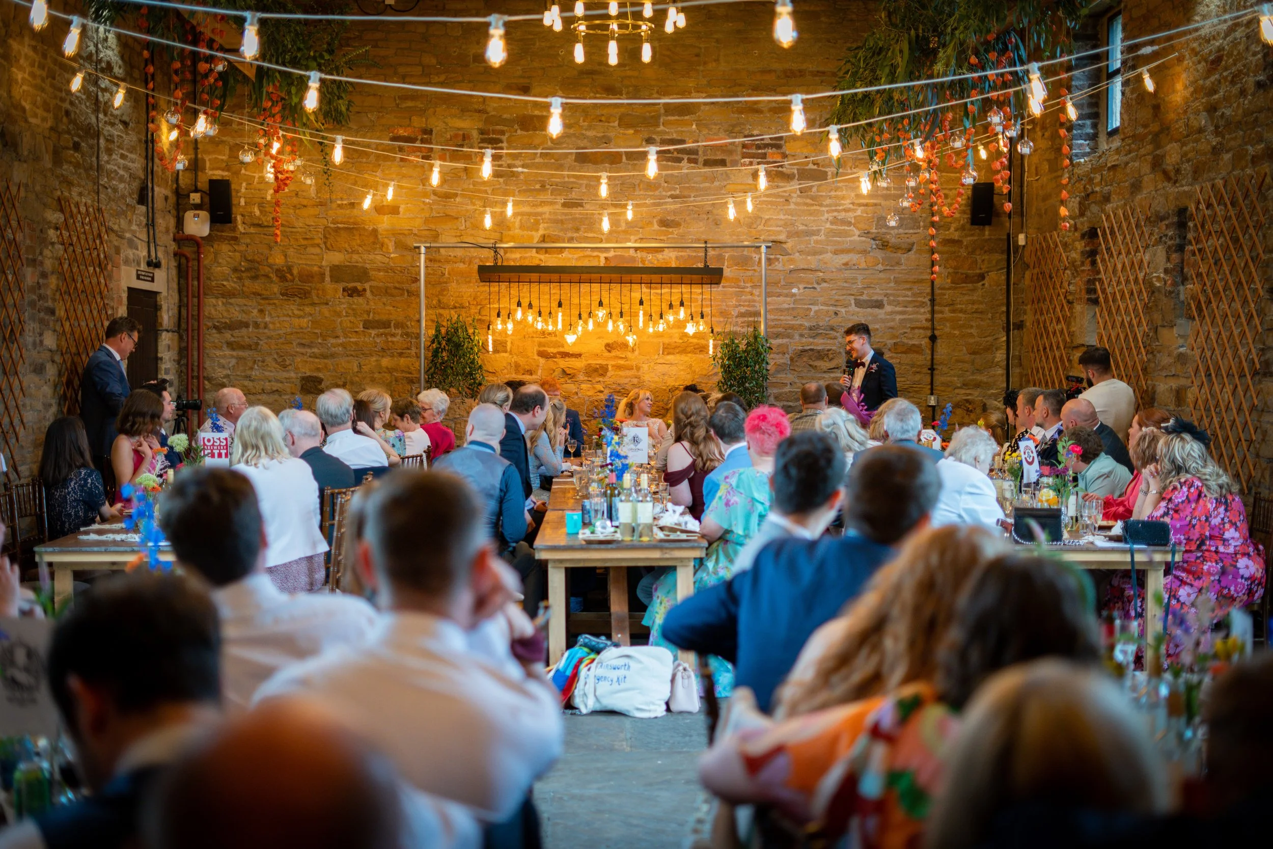 Wedding reception in a rustic indoor venue with exposed brick walls, decorated with hanging string lights and greenery, featuring many seated guests listening to a speaker.