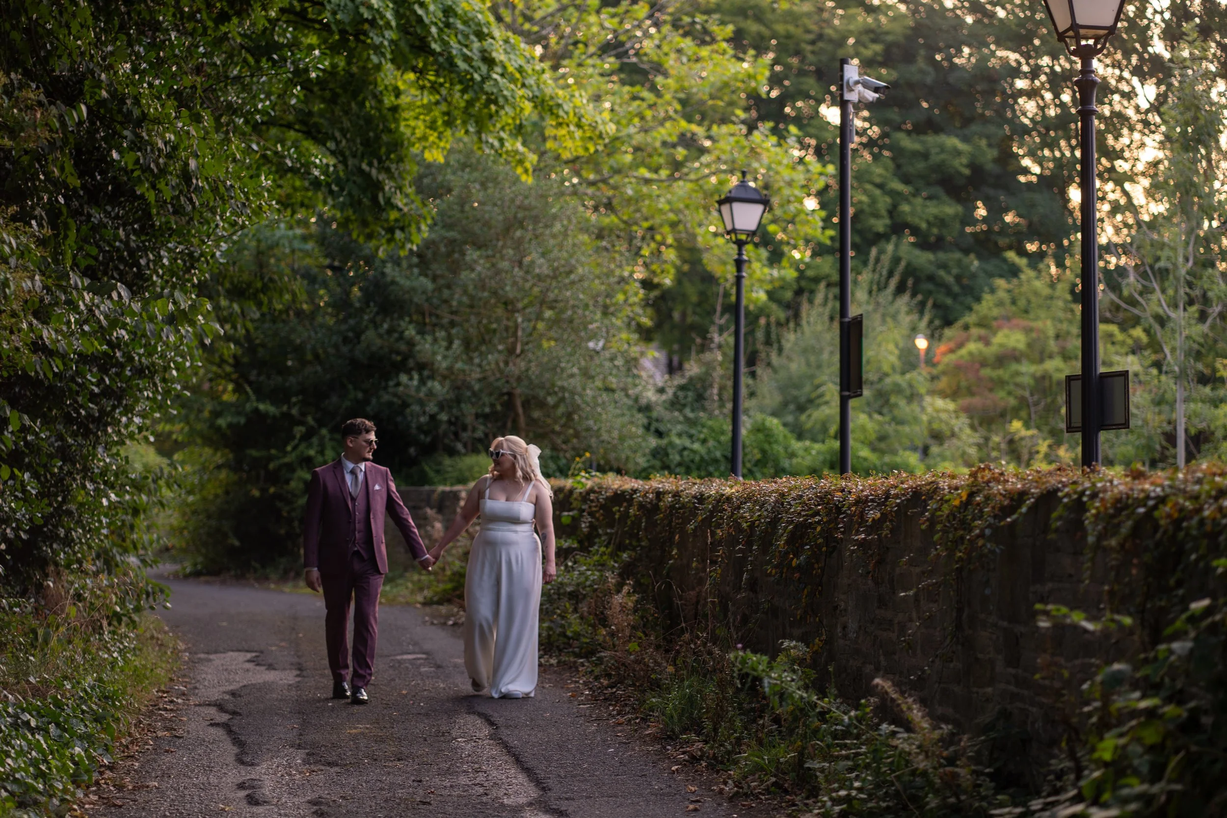 A couple in formal attire walking hand in hand on a tree-lined path with vintage street lamps during sunset.