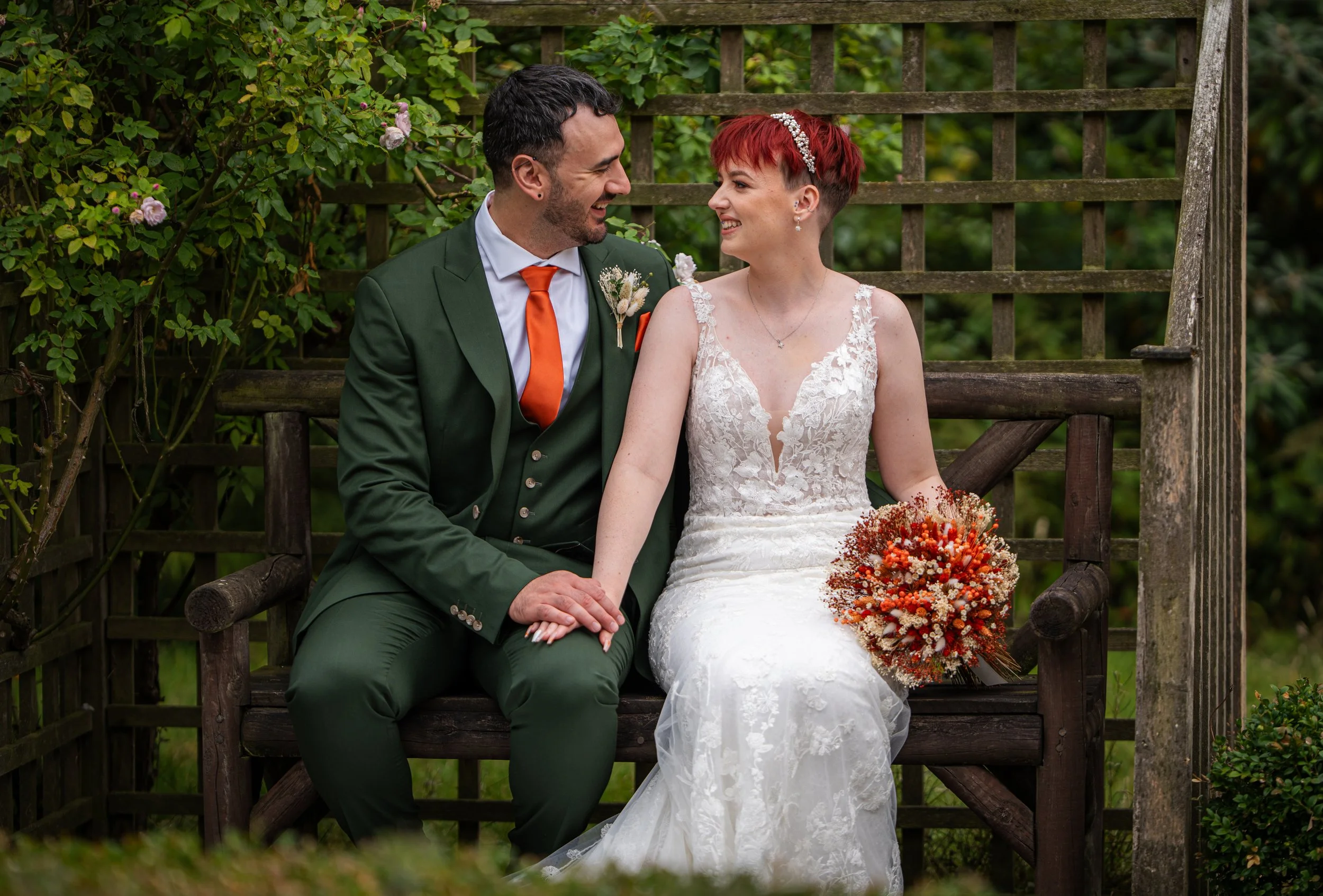 A newlywed couple sitting on a wooden bench in a garden, smiling at each other. The groom is wearing a green suit with an orange tie, and the bride is in a lace wedding dress with a bouquet of orange and red flowers.