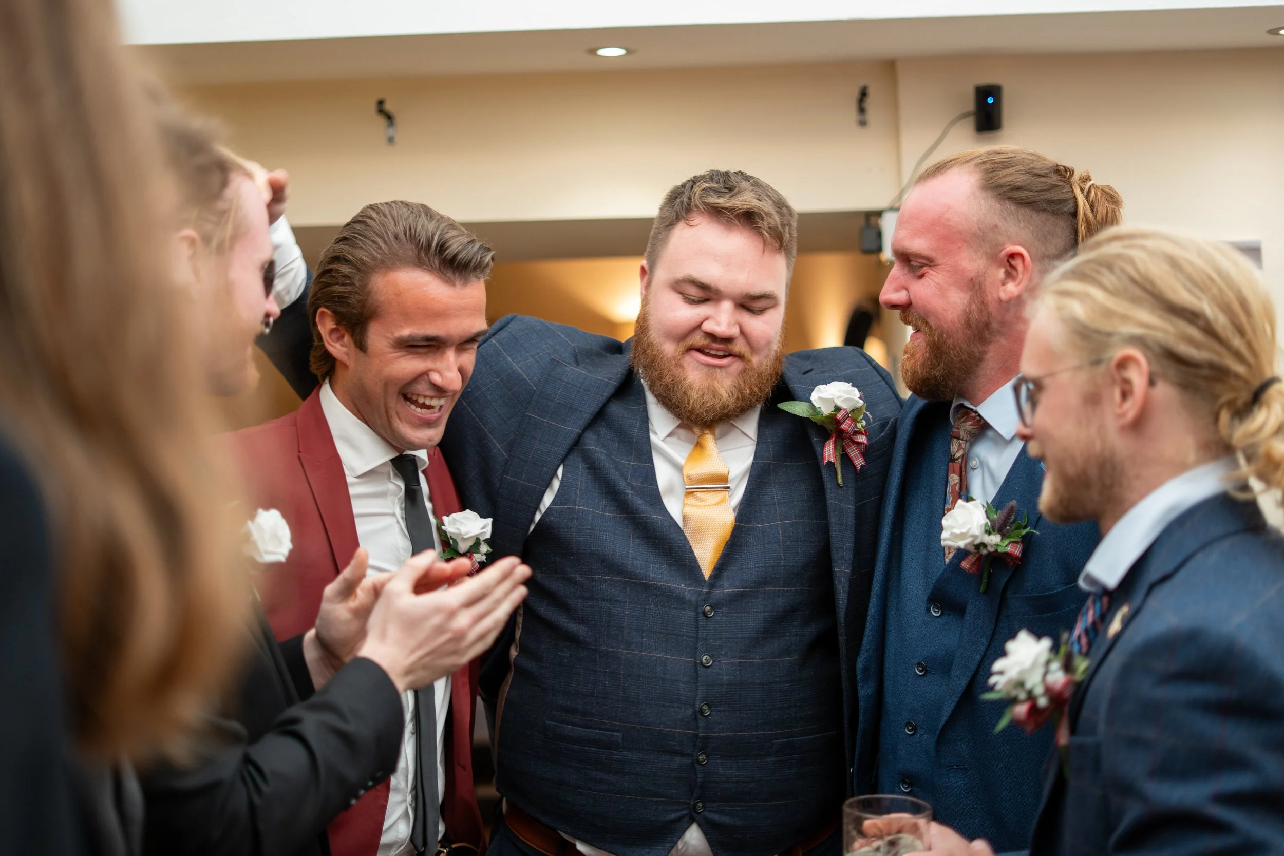 Group of men in suits with boutonnieres, smiling and celebrating at a social event.