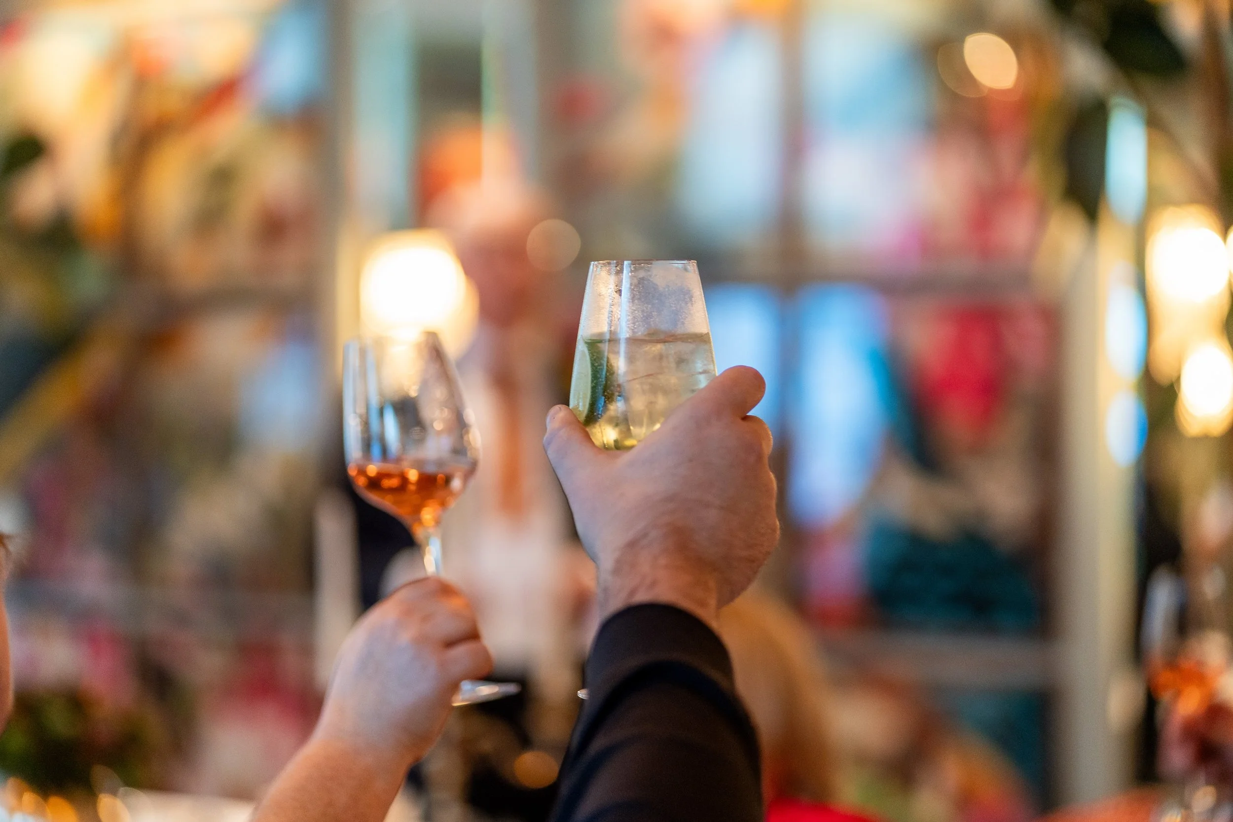 Two people holding glasses of drinks at a celebration or party, with colorful, blurred background lights and decorations.