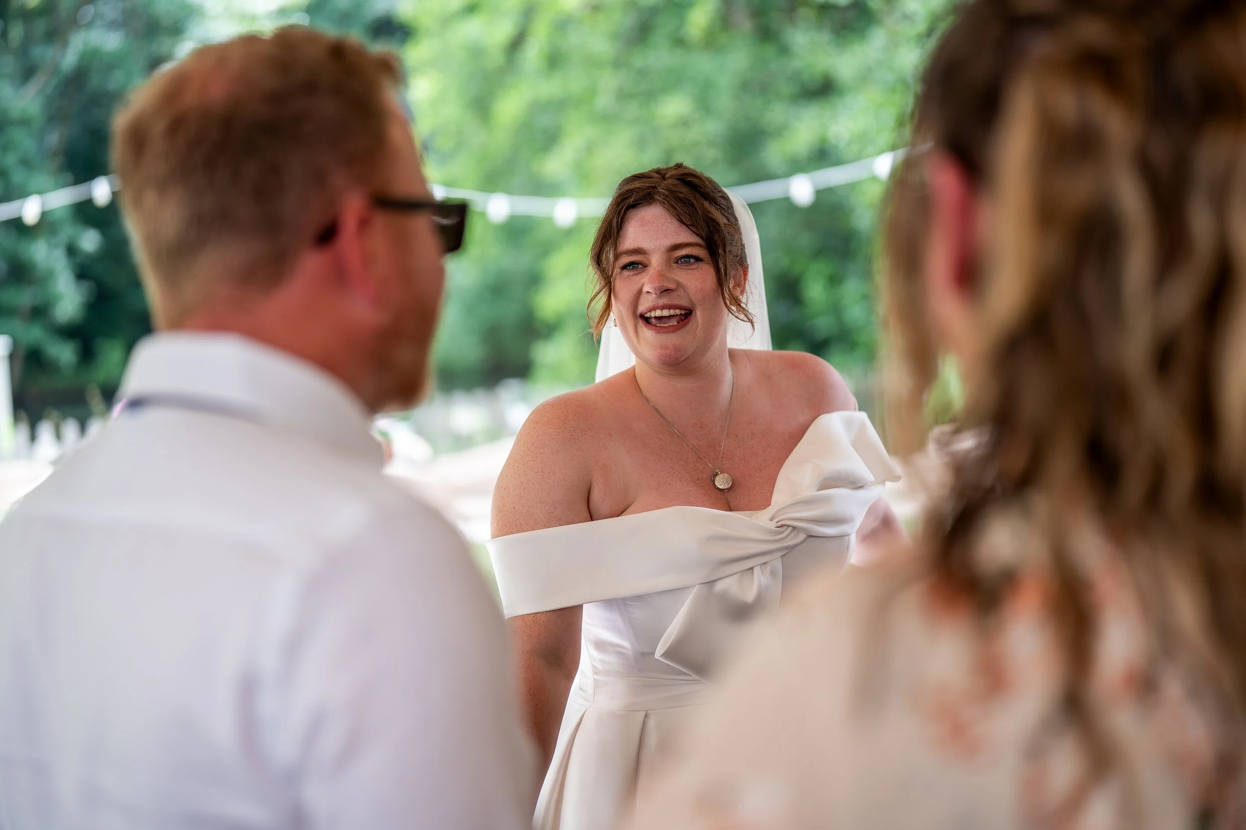A woman in a white dress with a bow on her shoulders smiling and talking to two men at an outdoor wedding reception, with trees and string lights in the background.