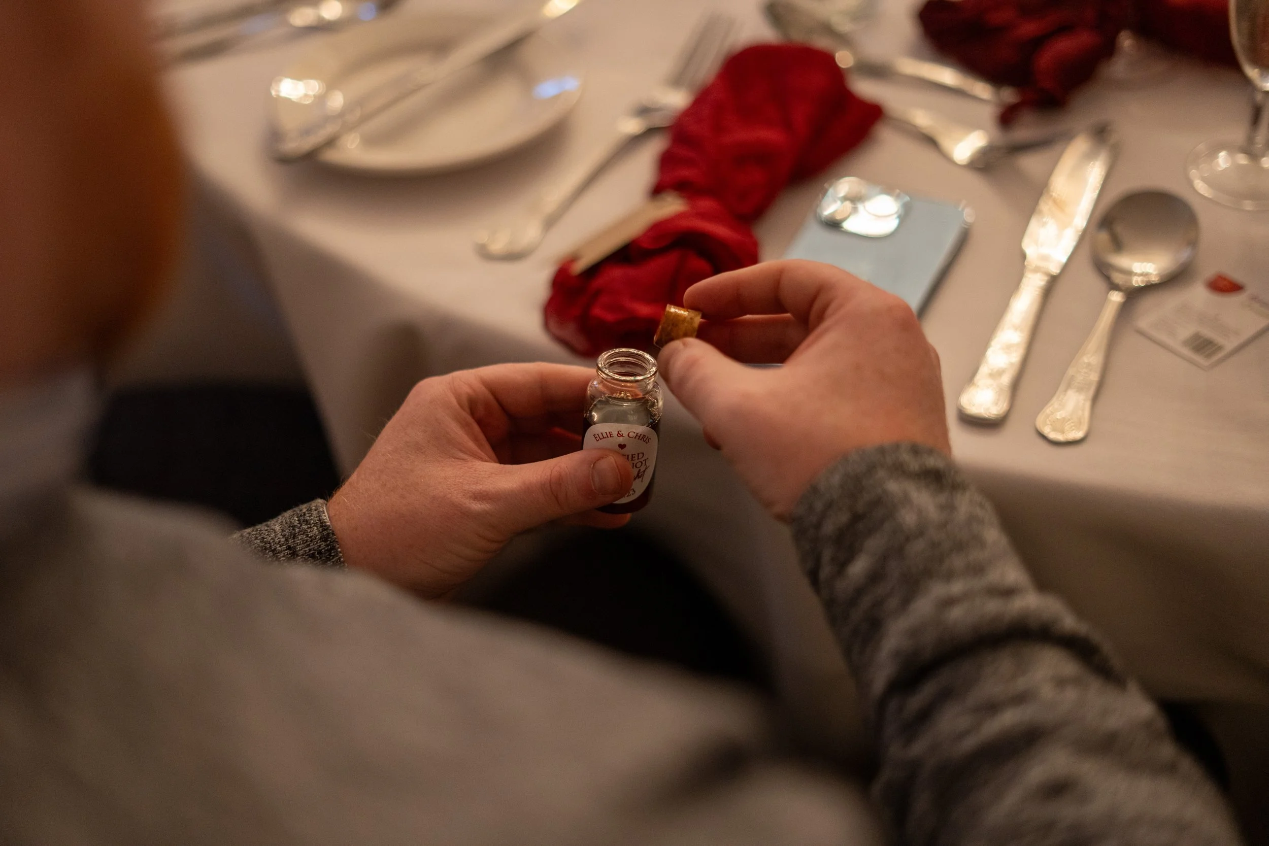 Person opening a small bottle of CBD oil at a dining table set with silverware, a smartphone, and a red napkin.