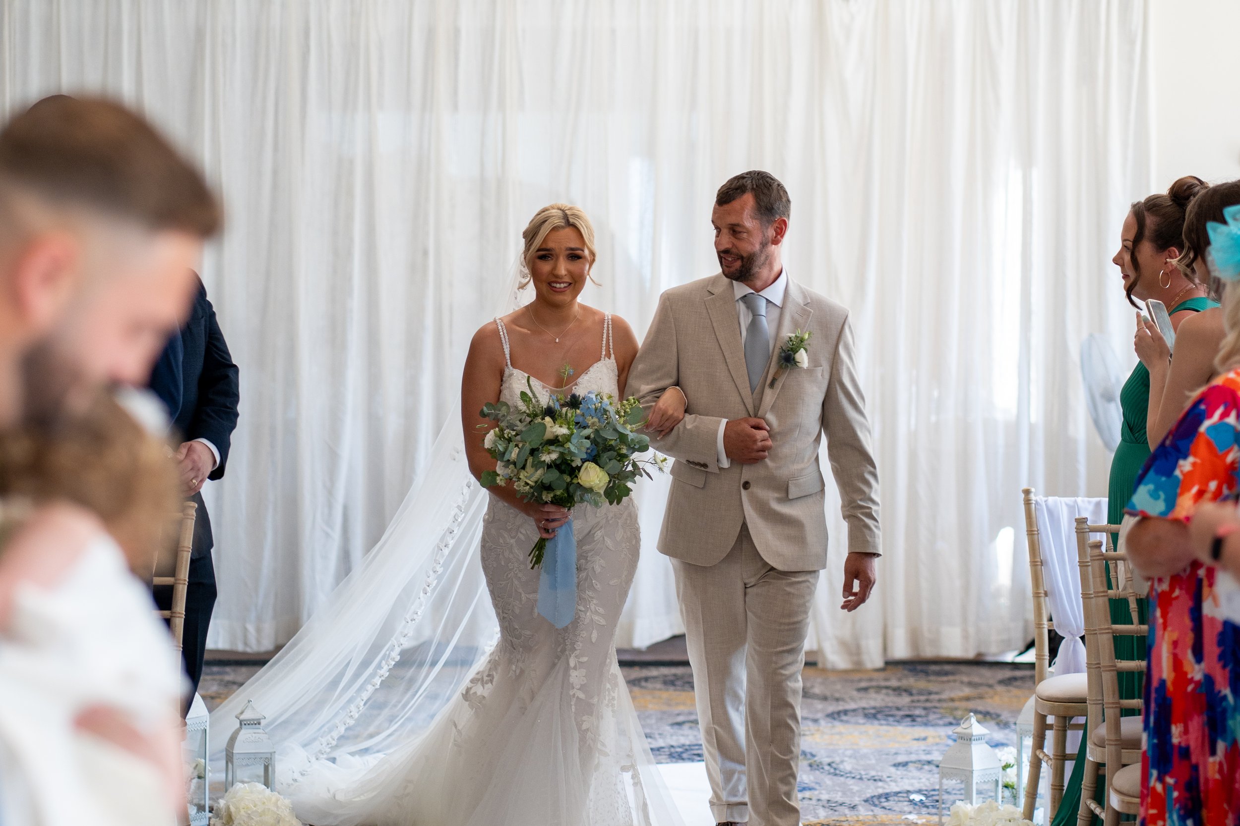 Bride walking down the aisle with her father during a wedding ceremony. The bride is holding a bouquet of white and blue flowers, and the venue has white drapery and chairs with white covers.