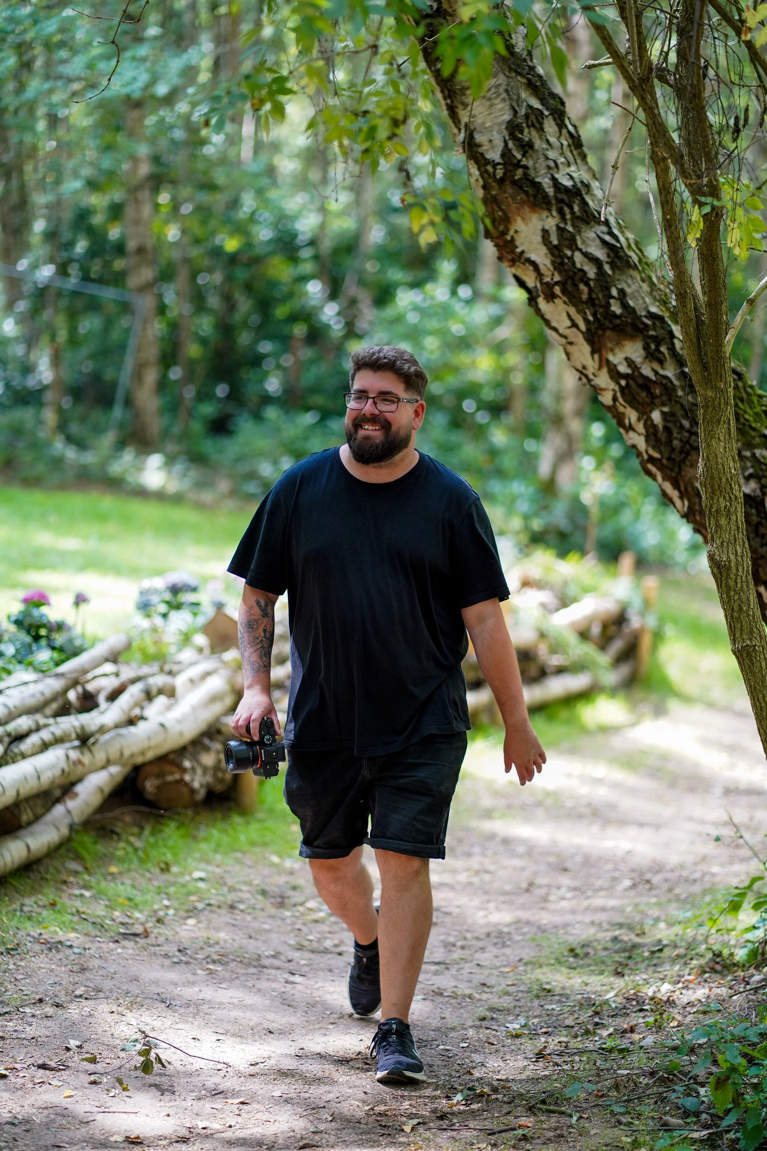A man with glasses and a beard walking along a dirt trail in a lush green forest holding a camera in his right hand. He is smiling and dressed in a black t-shirt and shorts, surrounded by trees and a fence made of logs.