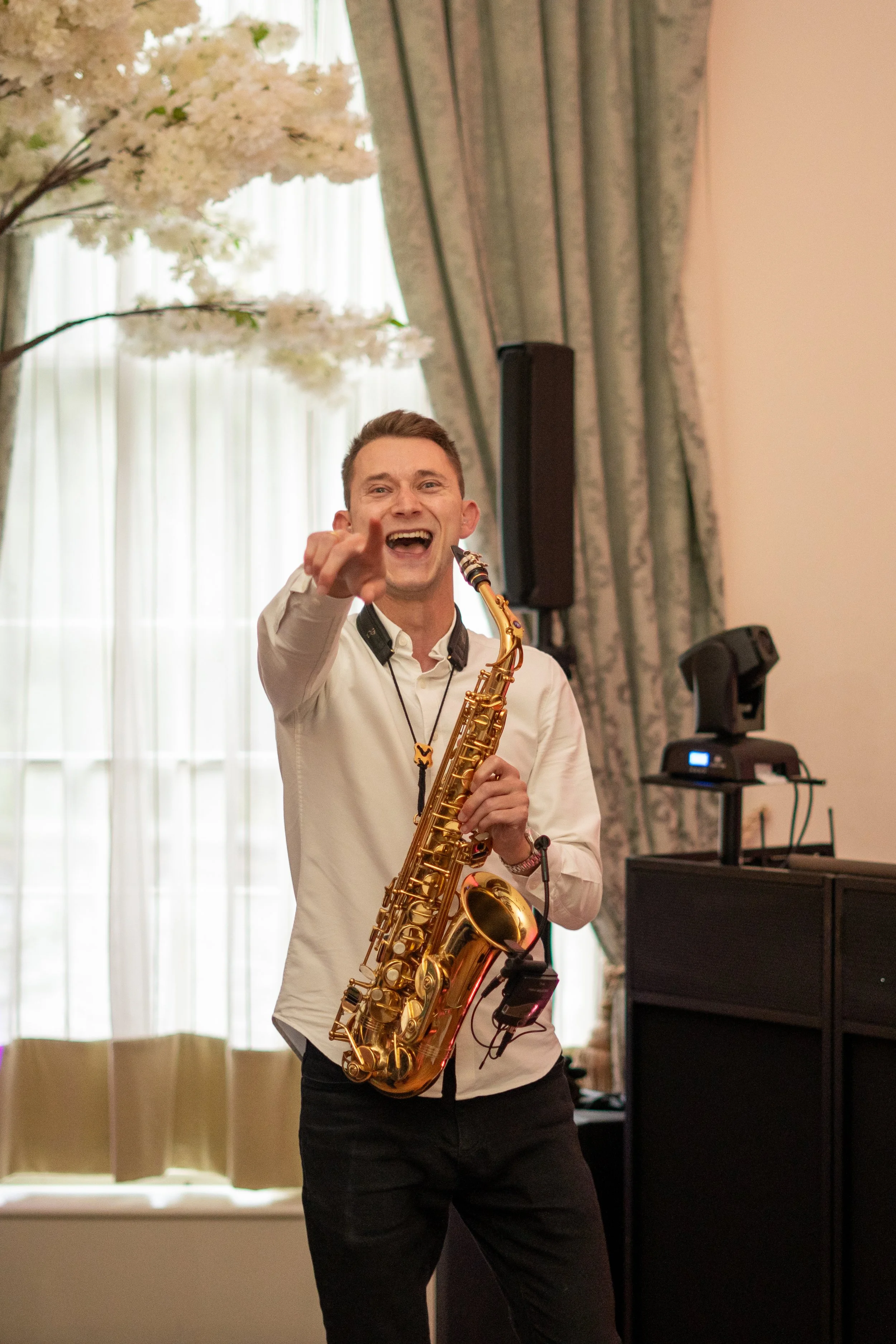 A man holding a saxophone, smiling, and pointing towards the camera in a bright room with floral curtains and a decorative tree.