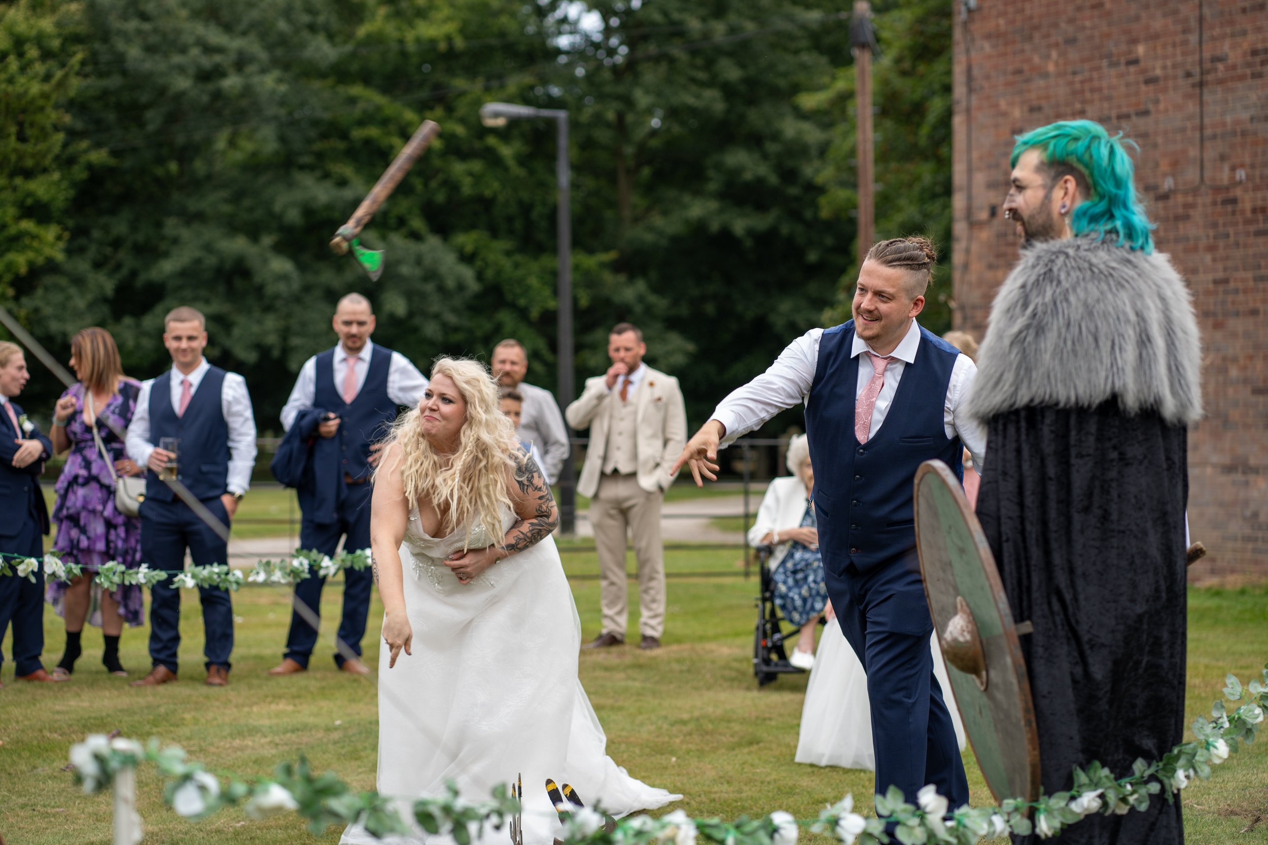 A wedding scene outdoors with a bride and groom participating in a traditional game, surrounded by guests in formal attire. The bride is squatting with a playful expression, and the groom is reaching out to her. An officiant or performer with blue ha