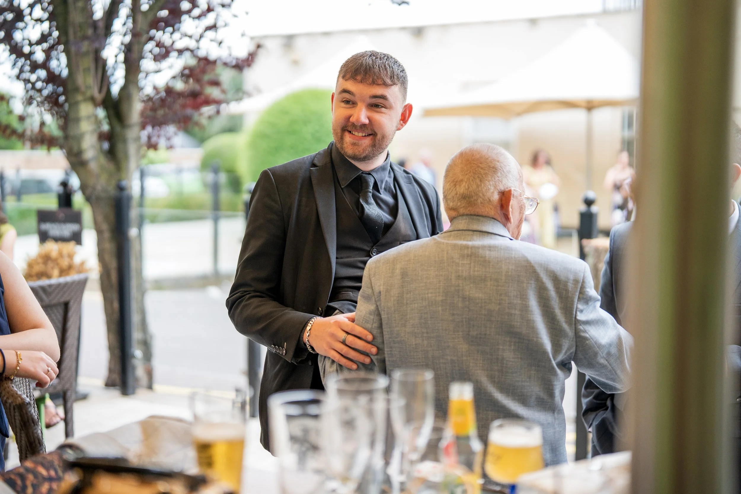 A young man in a black suit smiling and standing next to an older man in a gray suit at an outdoor event.