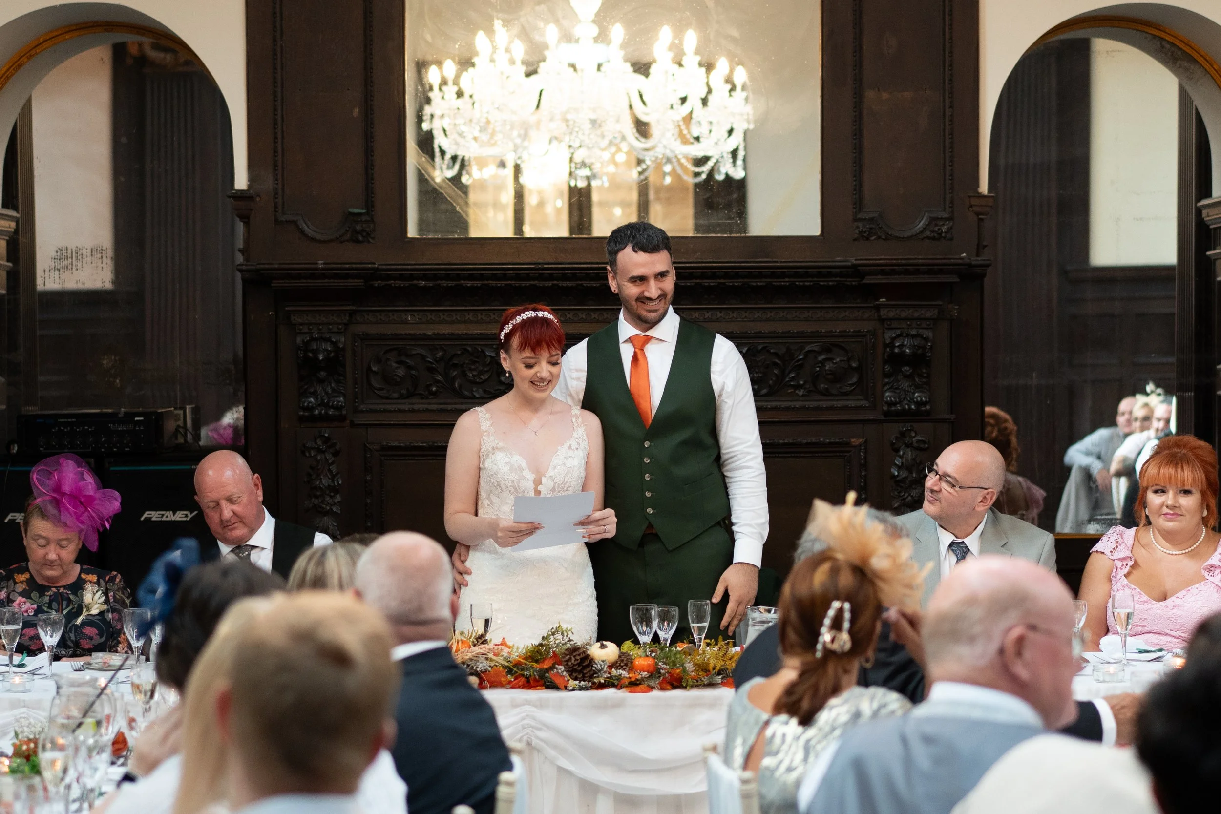 A bride and groom standing together at their wedding reception, smiling. The bride holds a paper, and guests are seated at tables around them. A chandelier is visible in the reflection behind them.