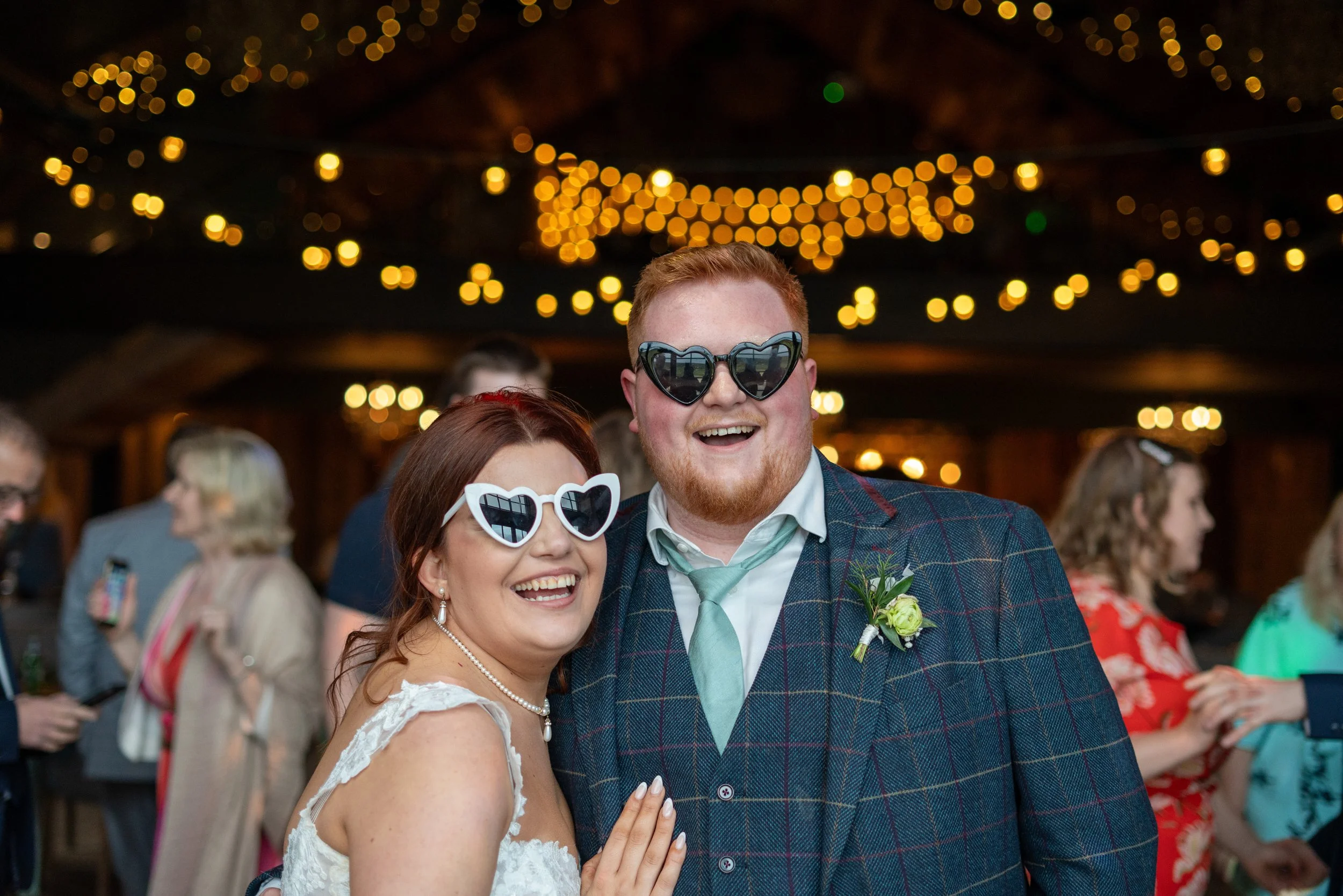 A couple at a wedding reception wearing heart-shaped sunglasses, smiling and having fun, with string lights in the background.