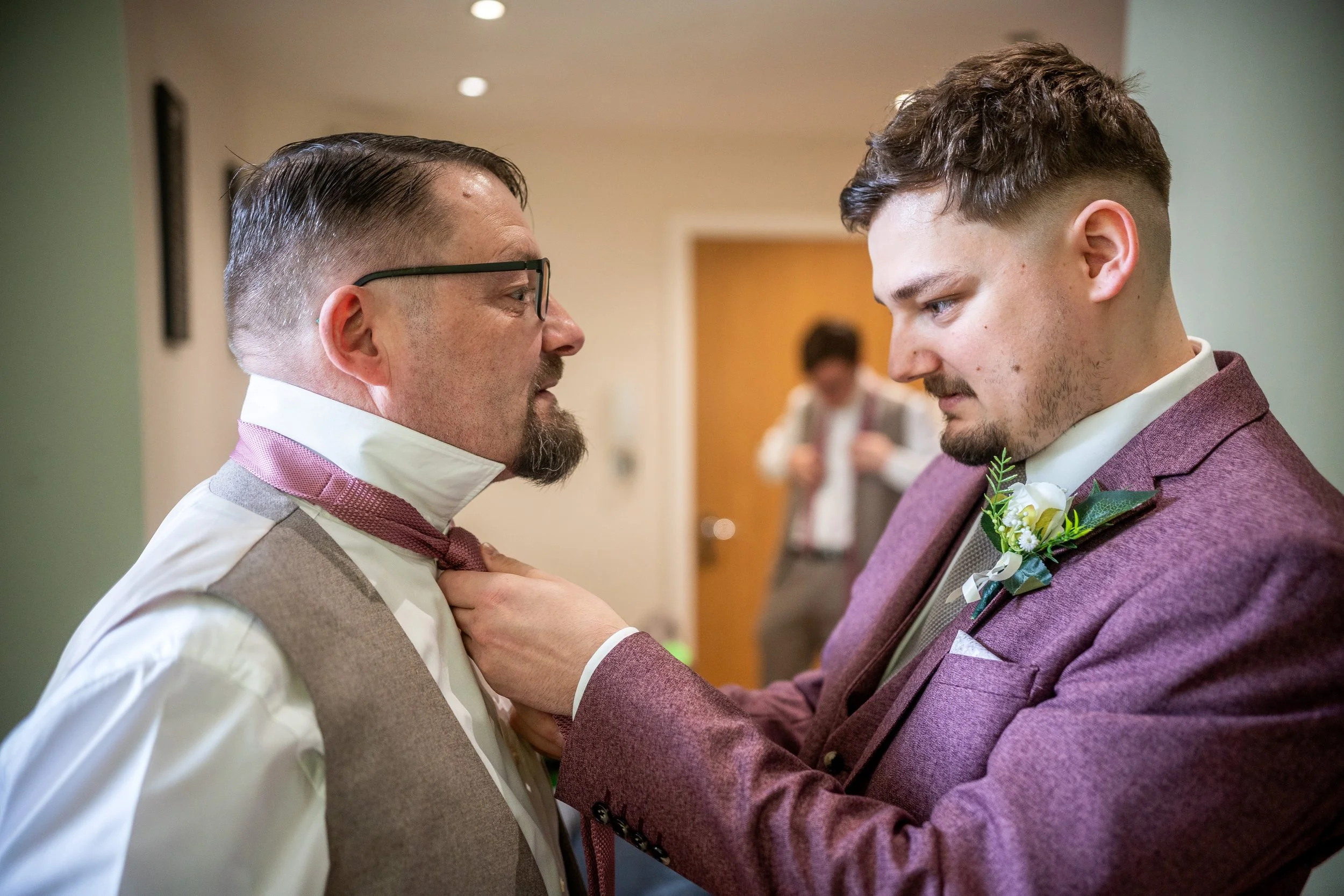 A man in a white shirt and gray vest adjusting his purple bowtie with the help of another man in a purple suit and a boutonniere, in an indoor setting, with a blurred person in the background.