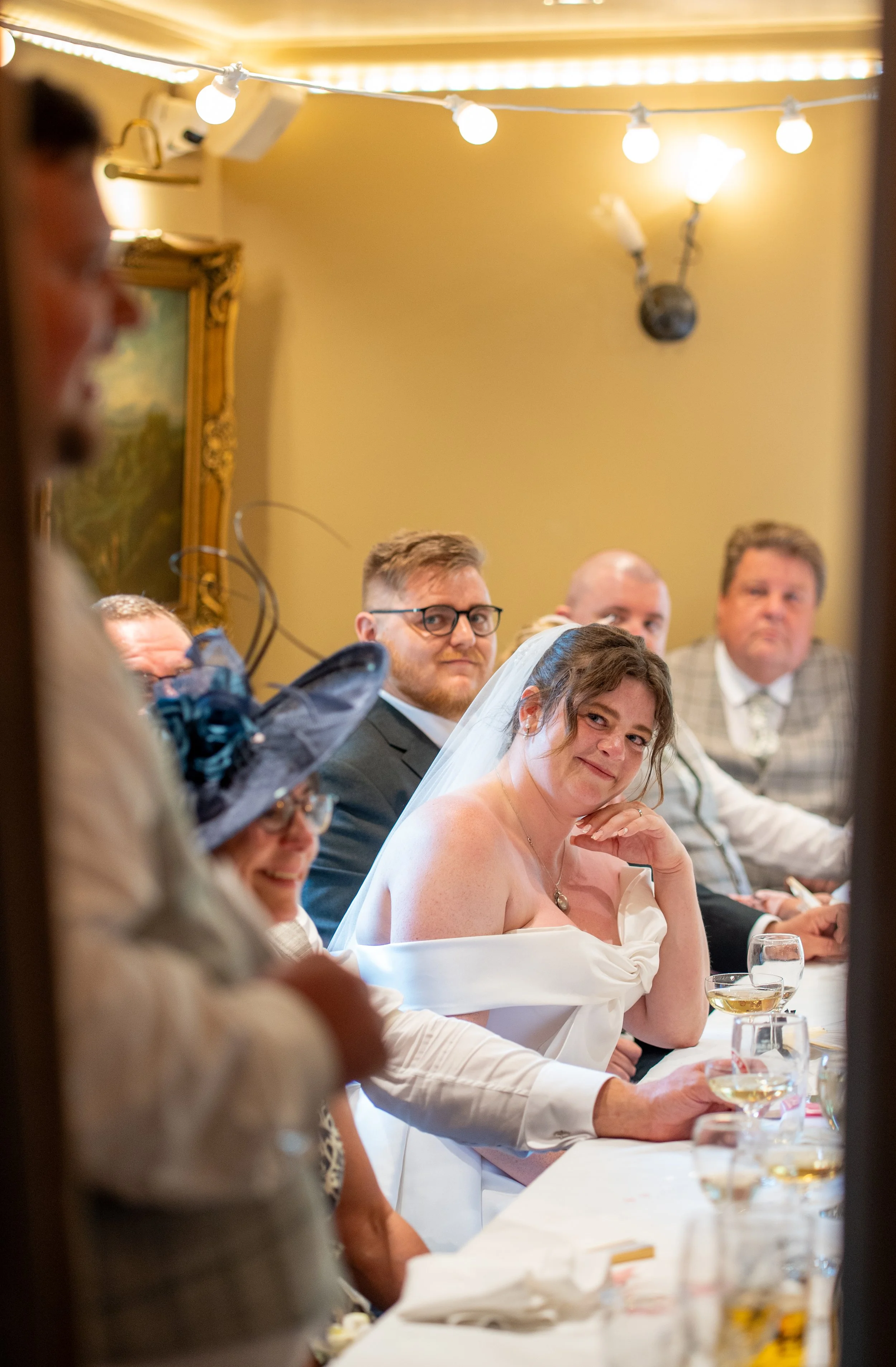 A bride sitting at a wedding reception table, surrounded by guests, with several wine glasses in front of her, smiling and looking at the camera.