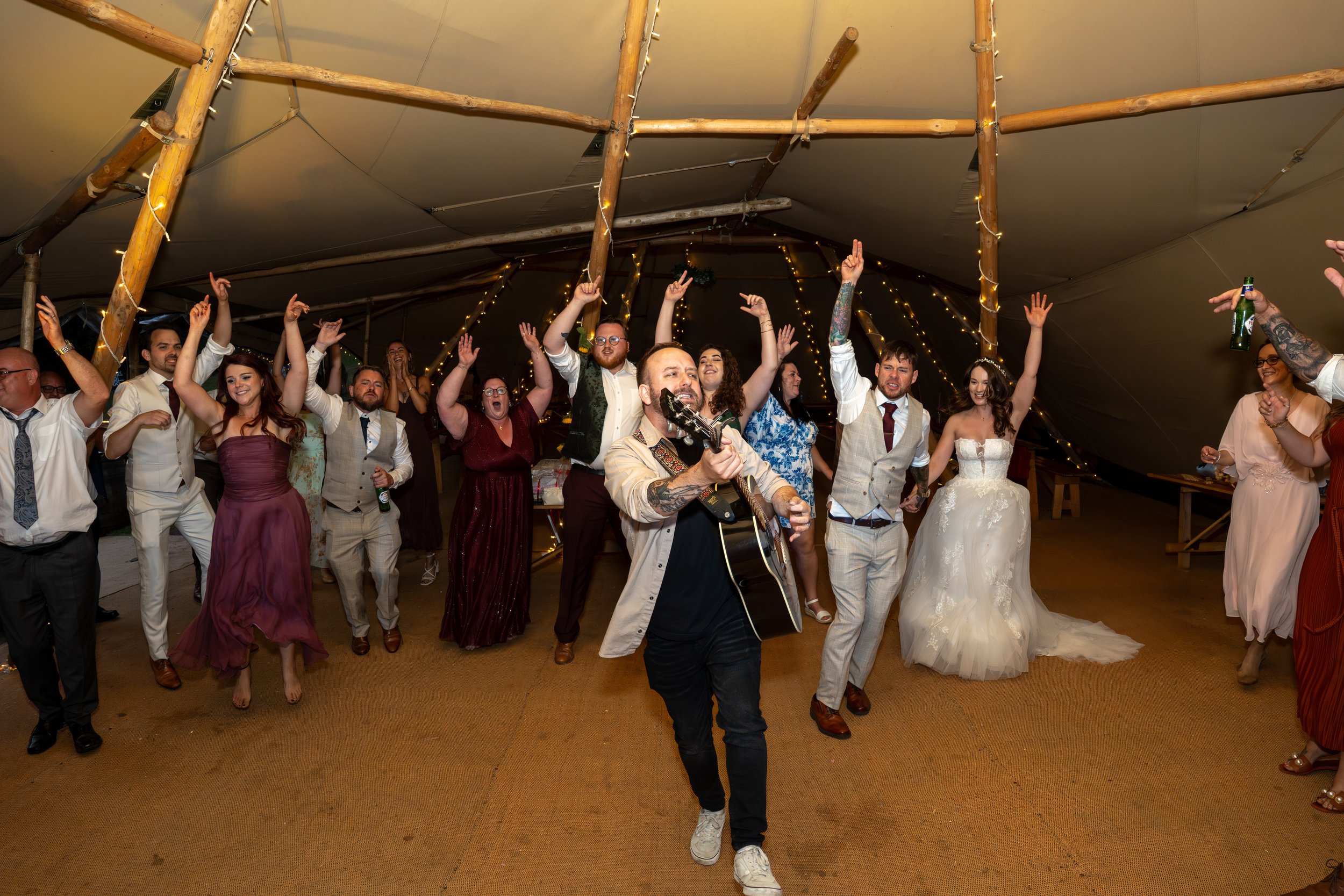 Group of people dancing and celebrating at a wedding reception with a live musician playing guitar in a decorated tent.