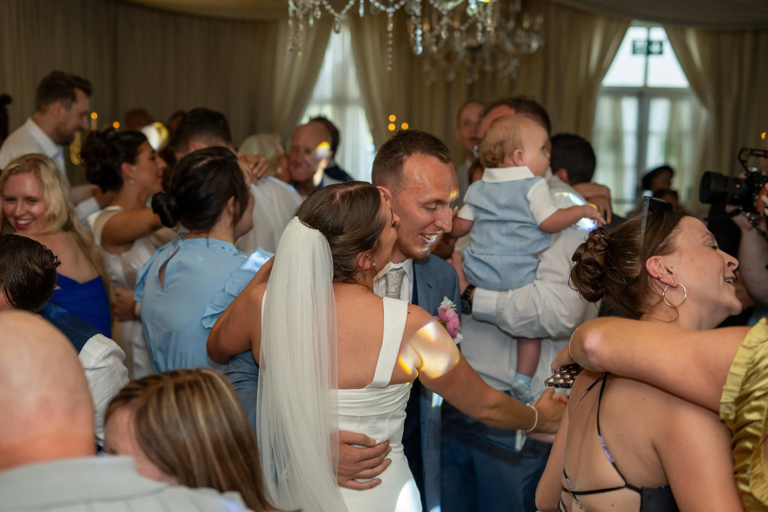 People dancing and celebrating at a wedding reception in an elegant indoor venue with chandeliers and large windows.