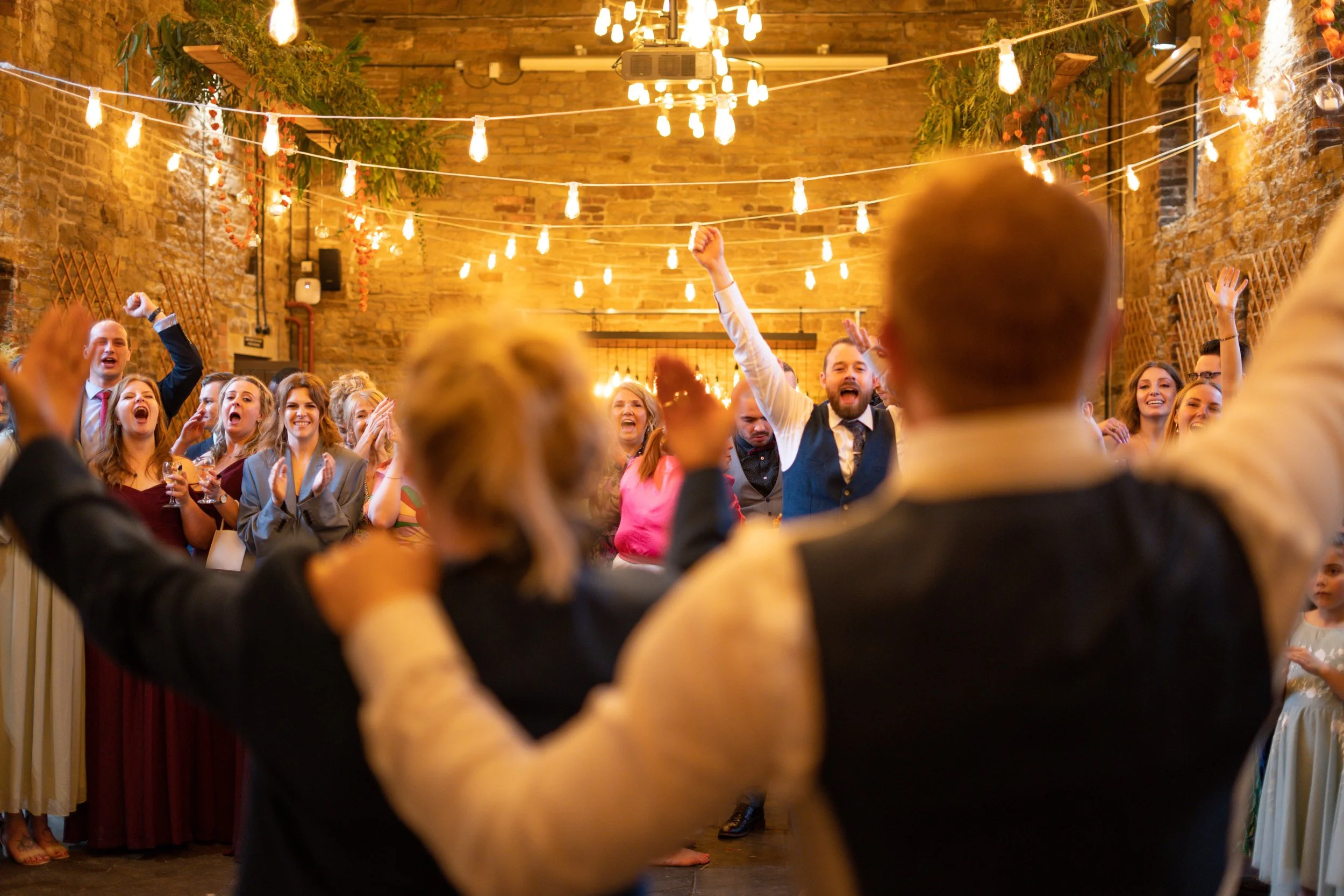 People celebrating and dancing at a wedding reception in a rustic venue with brick walls and string lights.