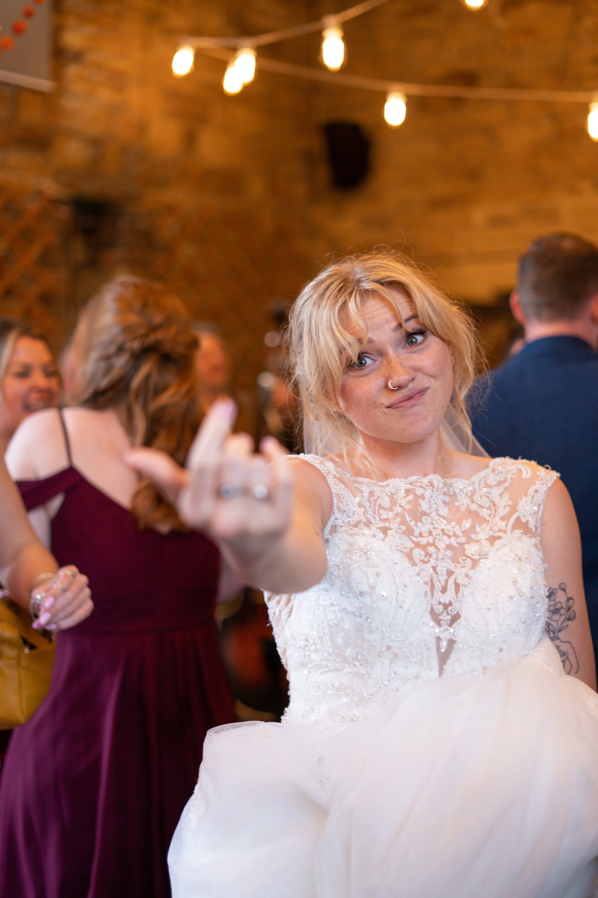 A woman in a white wedding dress making a rude gesture with her middle finger at a wedding reception. The background shows other guests and string lights.