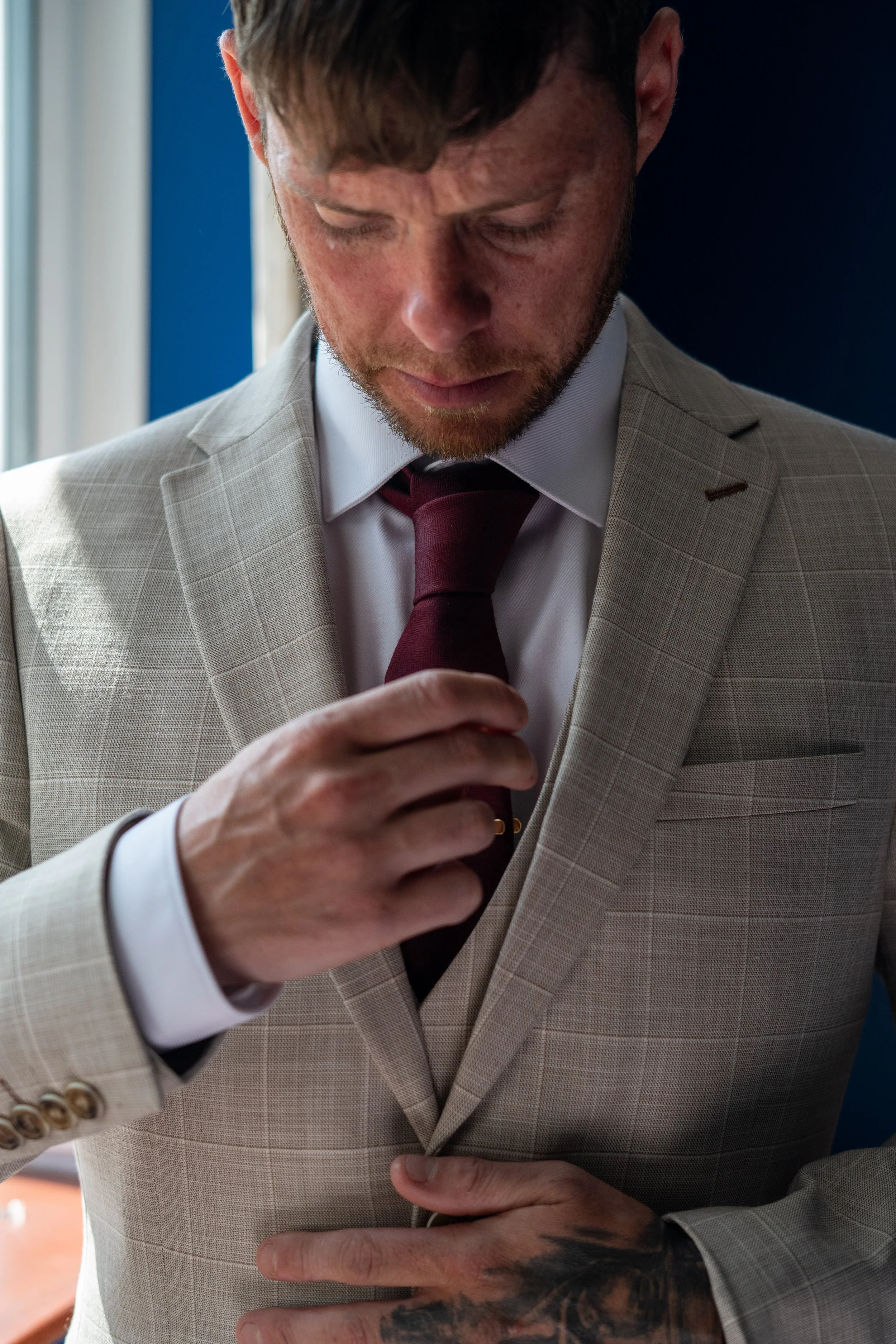 Man in beige suit adjusting maroon tie, with hand on buttoned jacket, standing indoors near window.