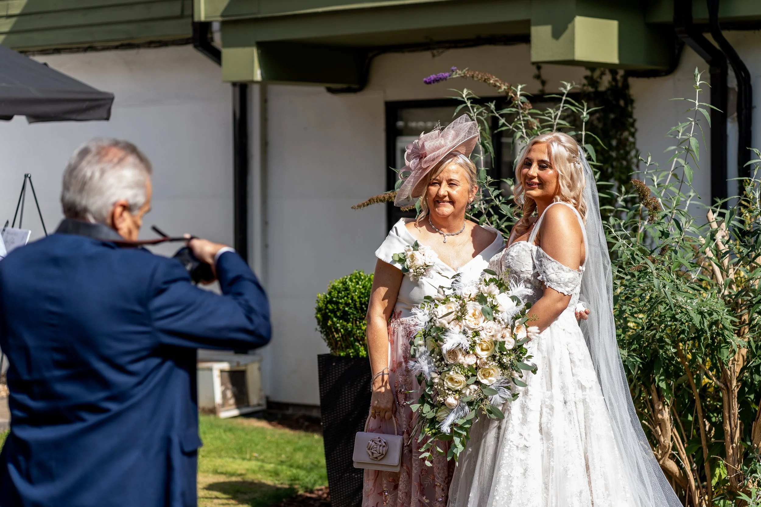 A bride in a white wedding dress and veil stands next to a woman in a pink hat and floral dress holding a large bouquet of flowers, while a man in a navy suit takes their photo outside on a sunny day.