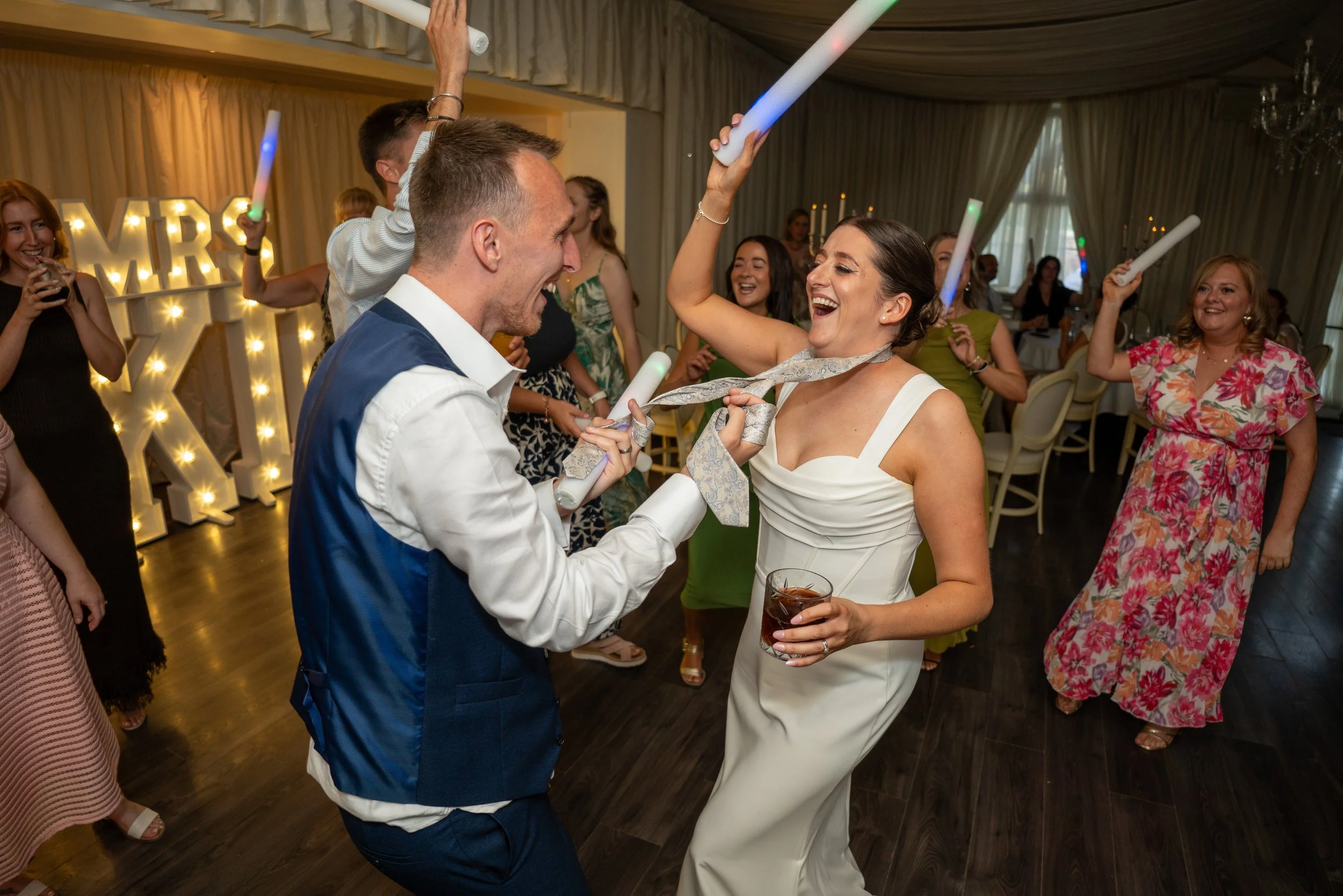 People celebrating at a wedding reception. A woman in a white dress is dancing and smiling with a drink in her hand, while a man in a white shirt and blue vest is joyfully interacting with her. Other guests are dancing and enjoying the celebration in