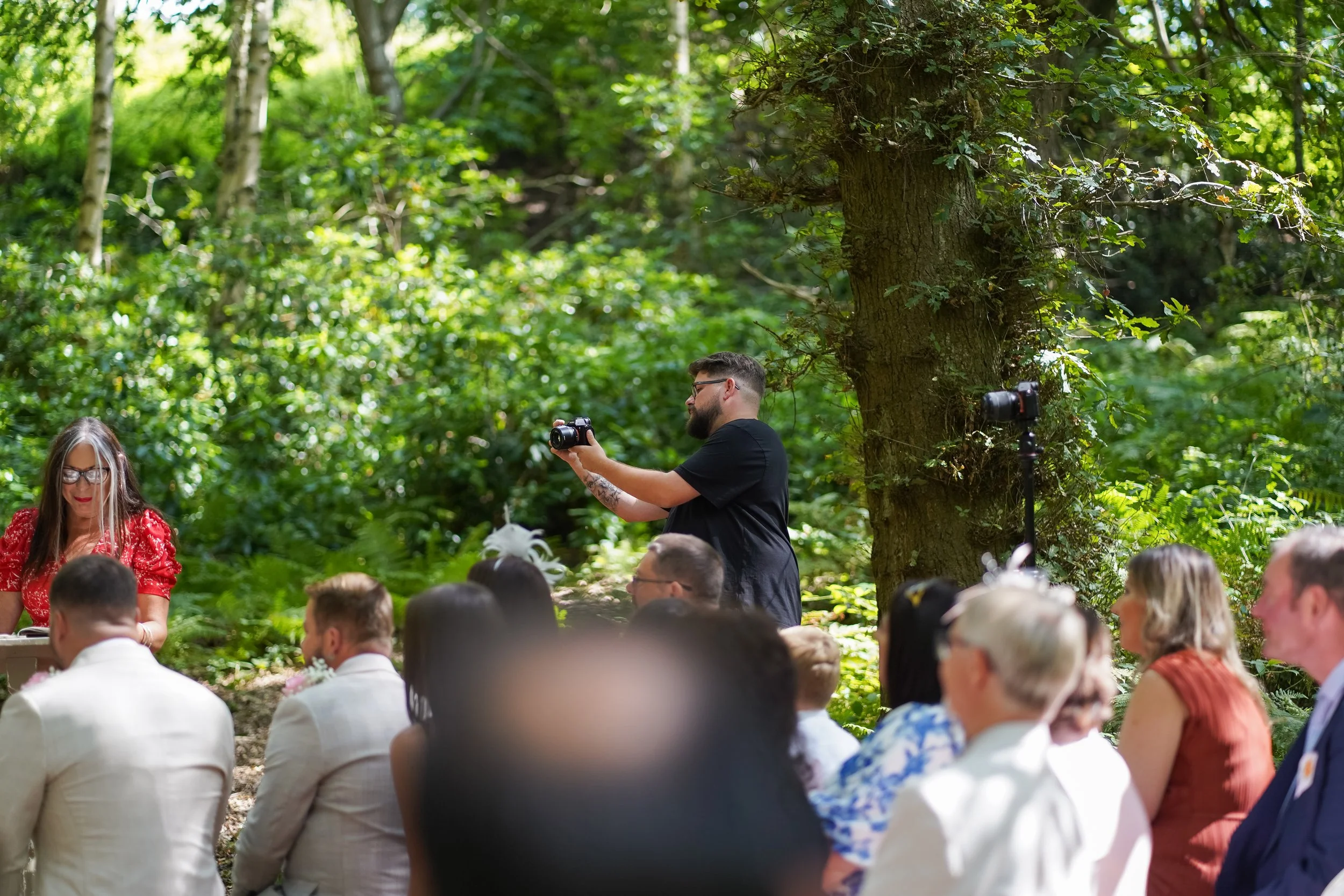 A wedding ceremony outdoors in a lush, green forest, with guests seated and a woman in a red dress standing nearby. A photographer in black shirt is taking photos, and there is a camera on a tripod.