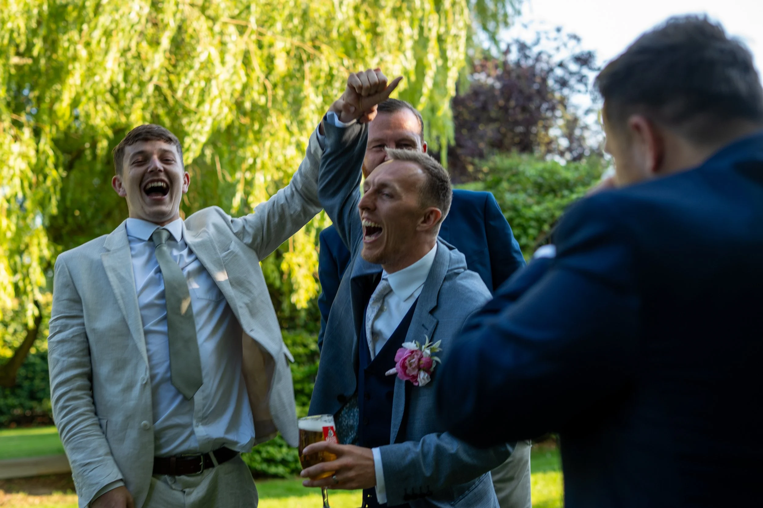 A group of men in suits celebrating outdoors with drinks and high-fives, surrounded by green trees.