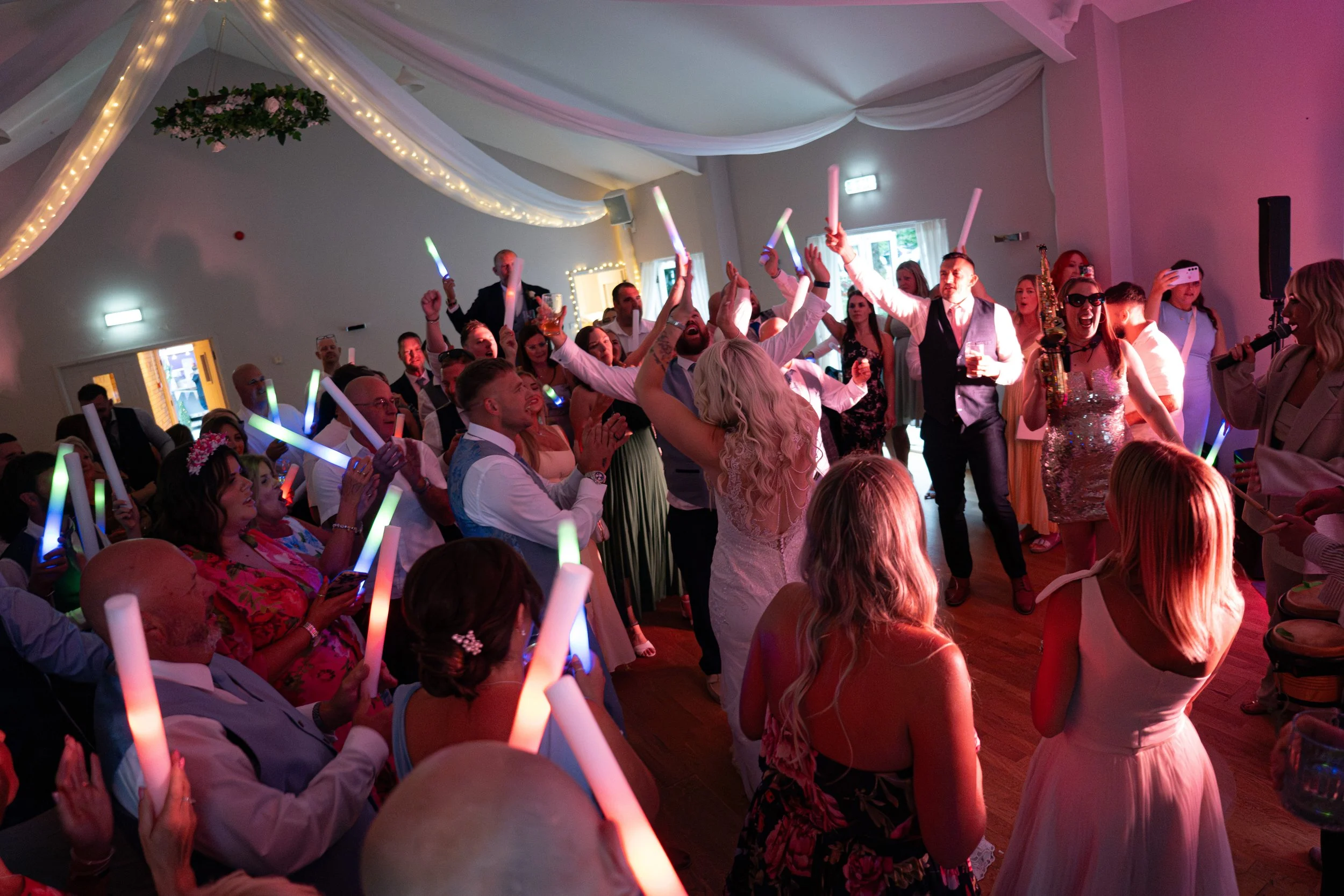 People dancing and celebrating at a wedding reception with colorful lights and glow sticks.