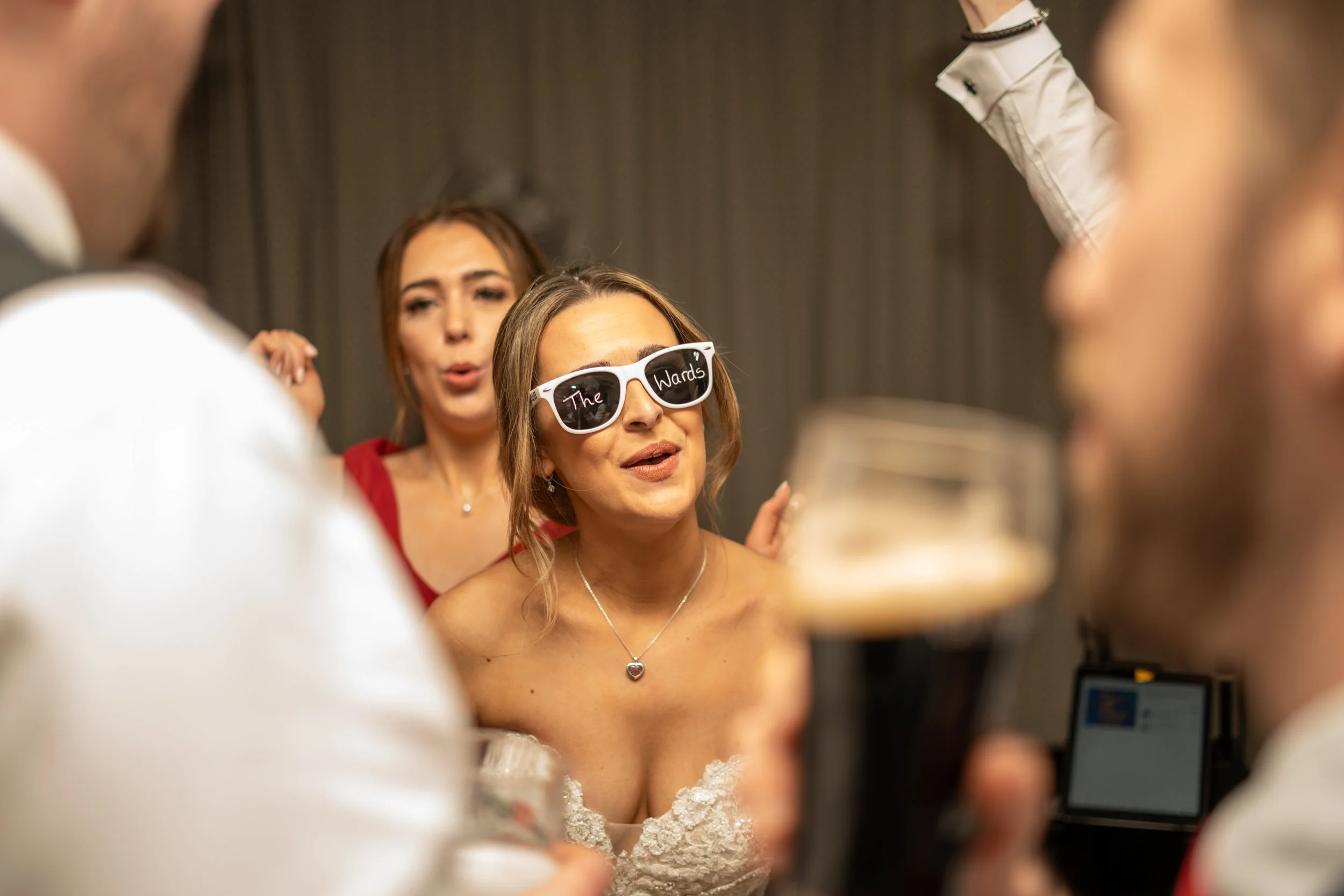 Women at a party, one wearing sunglasses with 'The Wards' written on them, speaking or singing, surrounded by people holding drinks in a dimly lit setting.
