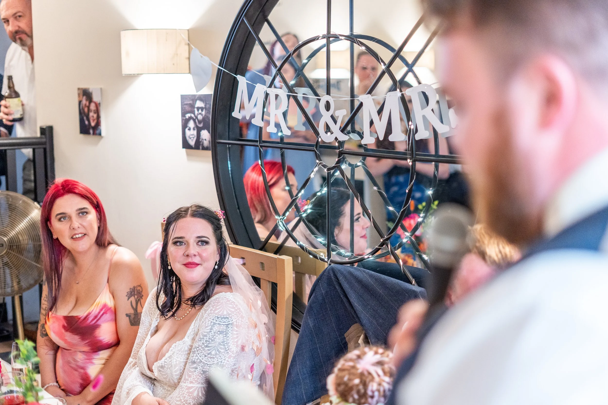 Happy wedding reception with two women sitting at a table, one with red hair and the other with black hair, smiling; a man in a tuxedo giving a speech in the foreground; 'MARRIAGE' decoration on a wall mirror in the background.