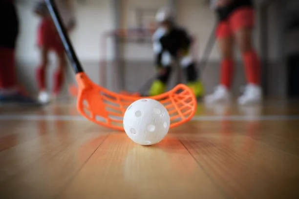 Floorball stick hitting a ball in an indoor gym, with players in the background.