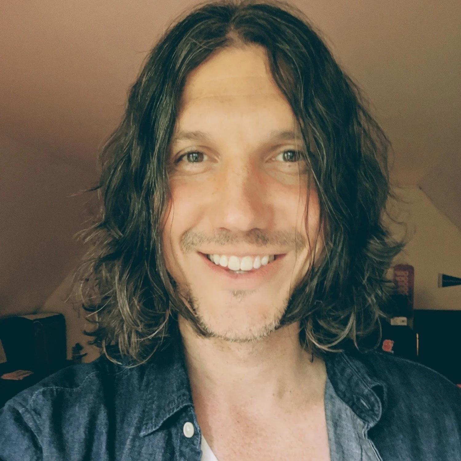 Smiling man with shoulder-length curly brown hair, wearing a dark denim shirt, indoors.