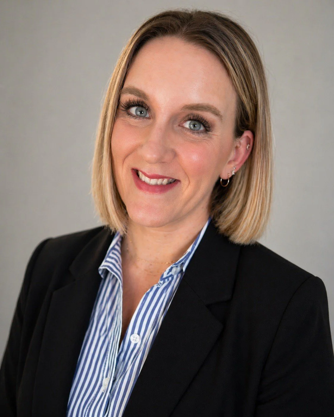 woman with blonde and brown bob, smiling wearing a black blazer and a striped denim and white button down top with gold necklaces