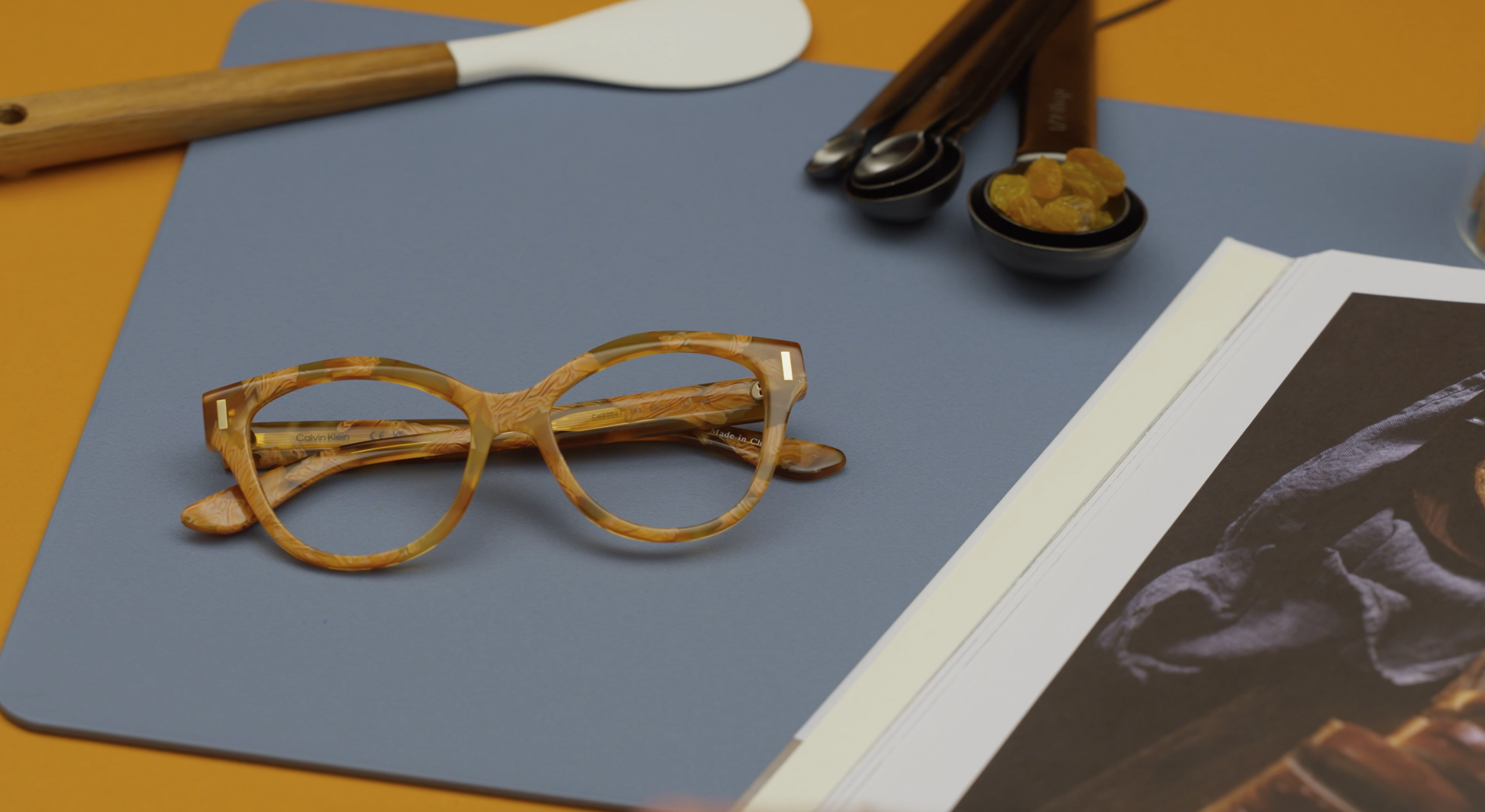 Calvin Klein camel or orange brown colored designed frames with blue base underneath, cooking book , spatulata wooden handle orangish background