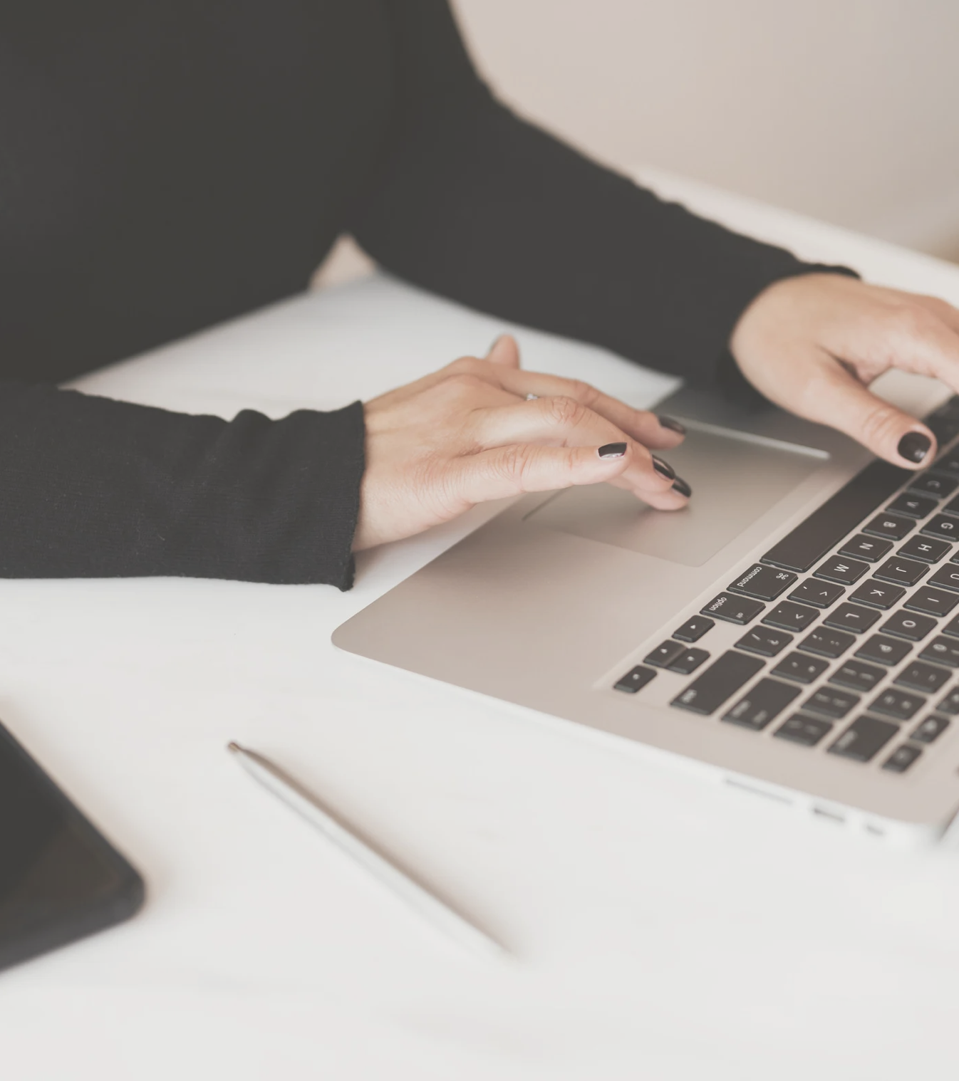 woman in black sweater at looks to be MacBook with black notebook by her, black nails typing on computer , black long sleeve shirt, grey pen white desk