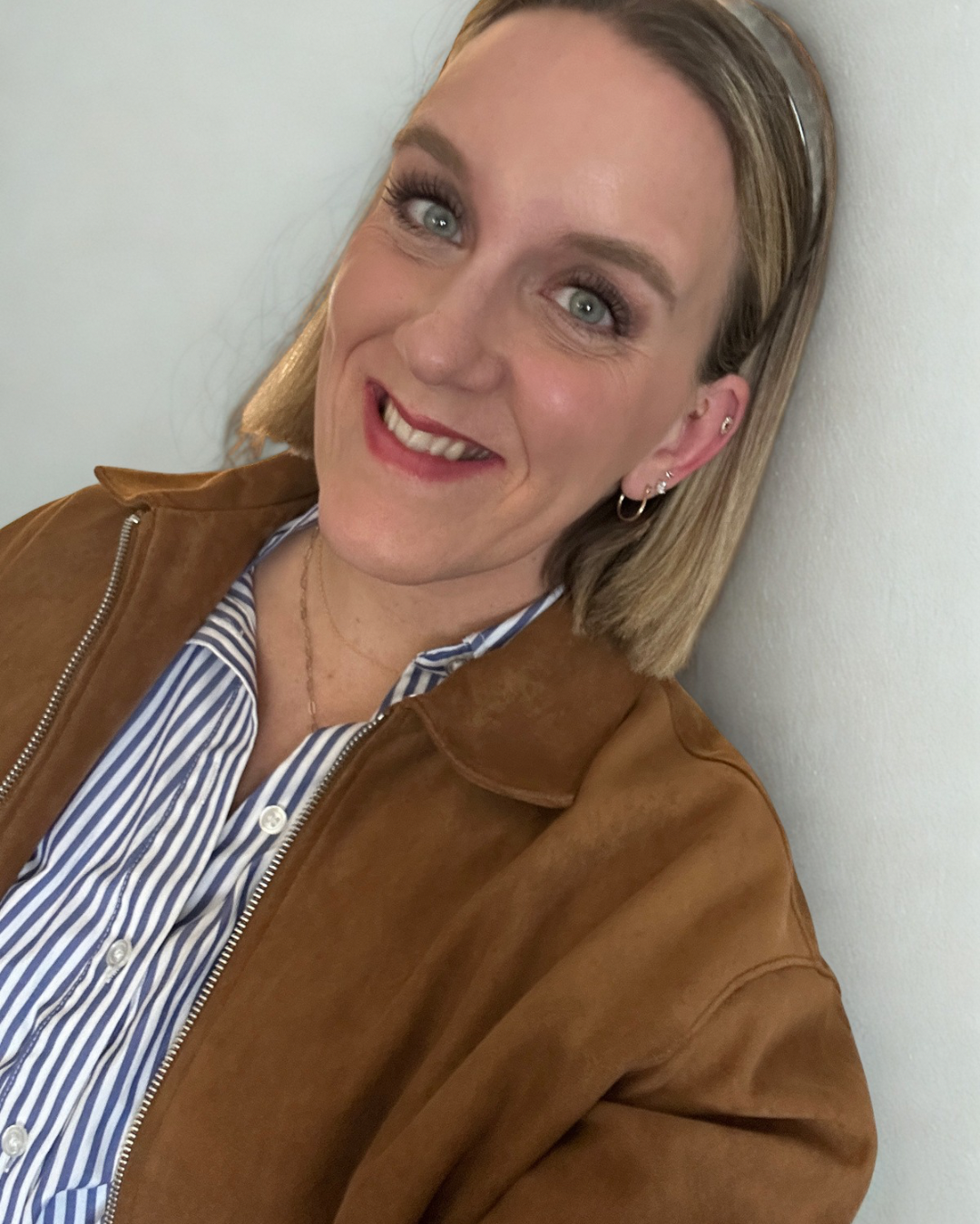 Woman short blonde bob, Carmel suede jacket headband in hai striped shirt cold jewelry blue green eyes with earrings, smiling.
