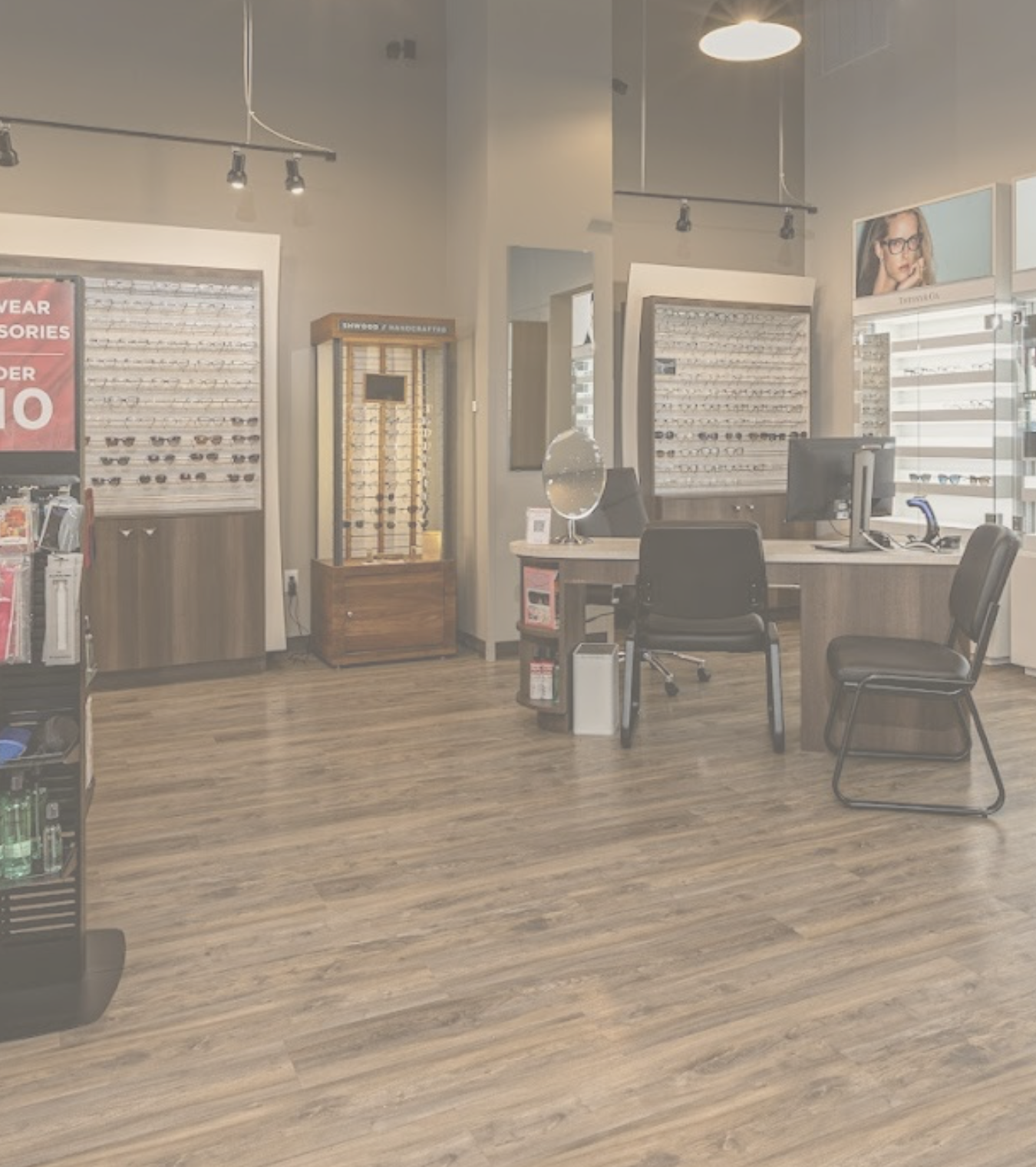 Inside an optical store with wooden floors, frame boards, chairs, mirror and computers, red sign partially shown with accessories, sunglasses and eyeglasses