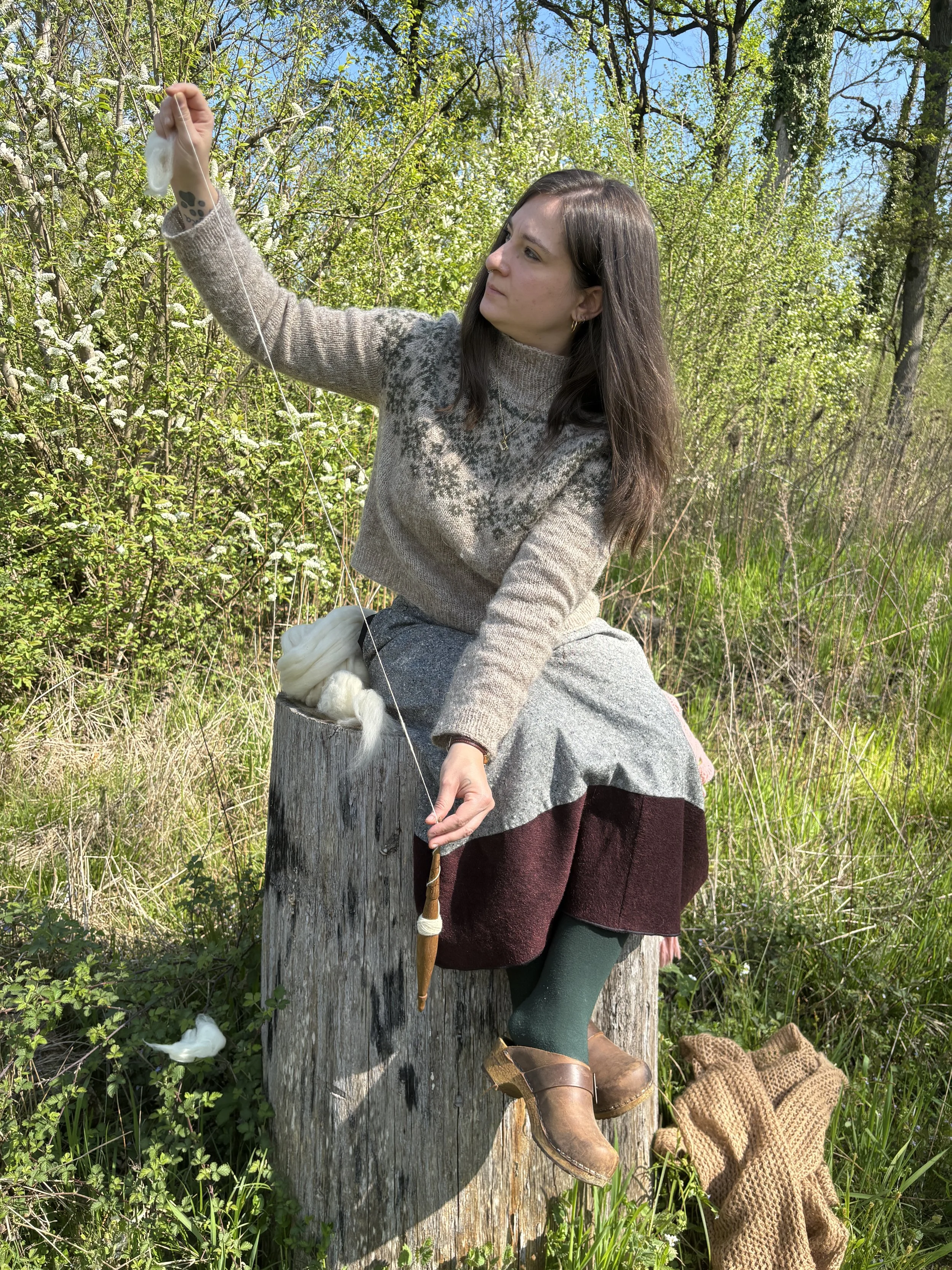Una donna con capelli castani e abbigliamento vintage siede su un tronco d'albero mentre tiene una bacchetta in mano e osserva attentamente un ramo. Sul prato vicino c'è un maglione di lana beige.