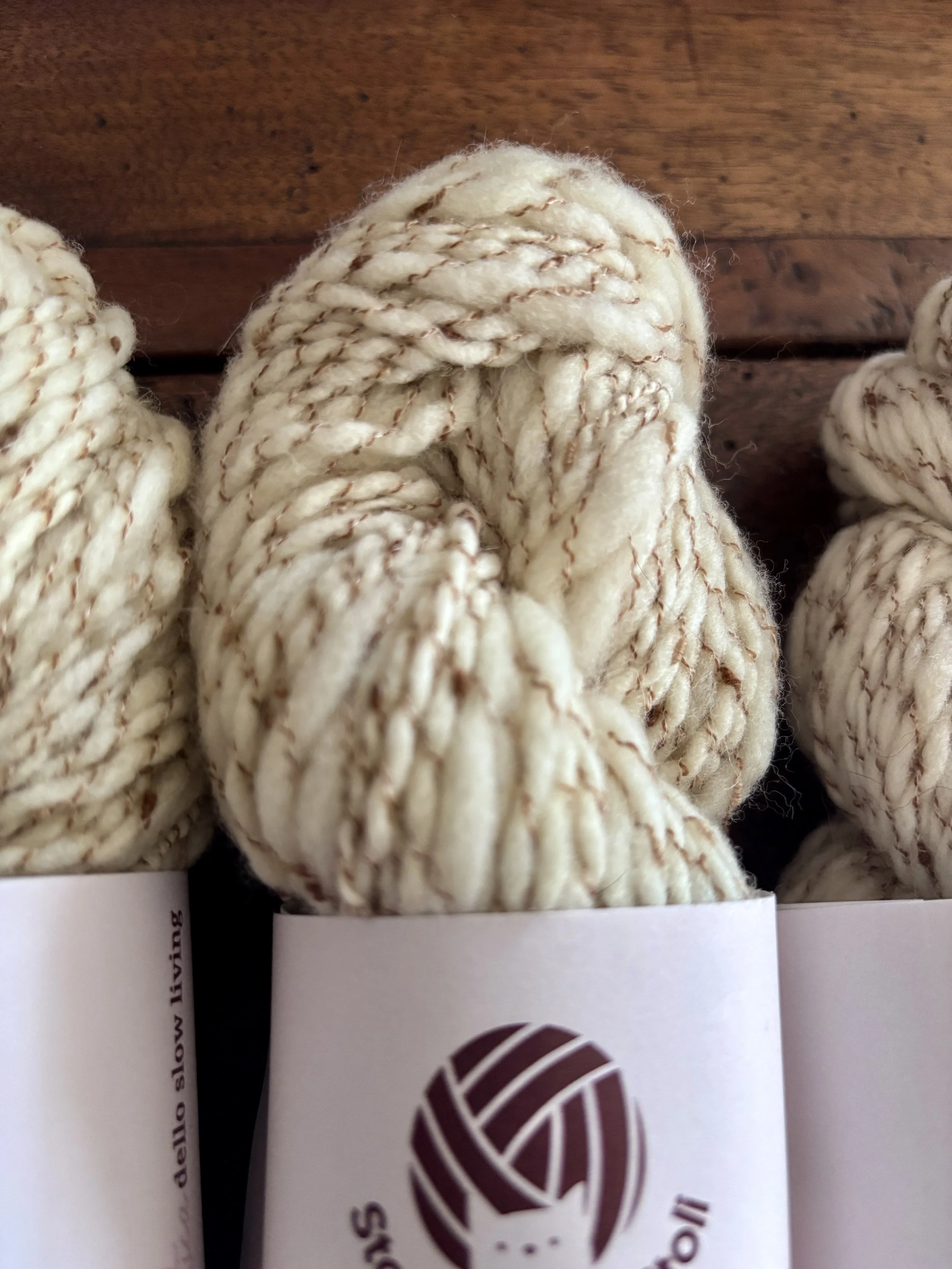 Palline di lana color crema con fili di colore marrone, in una confezione accanto a una scheda con logo stilizzato di un gomitolo e il testo 'Stili'.
