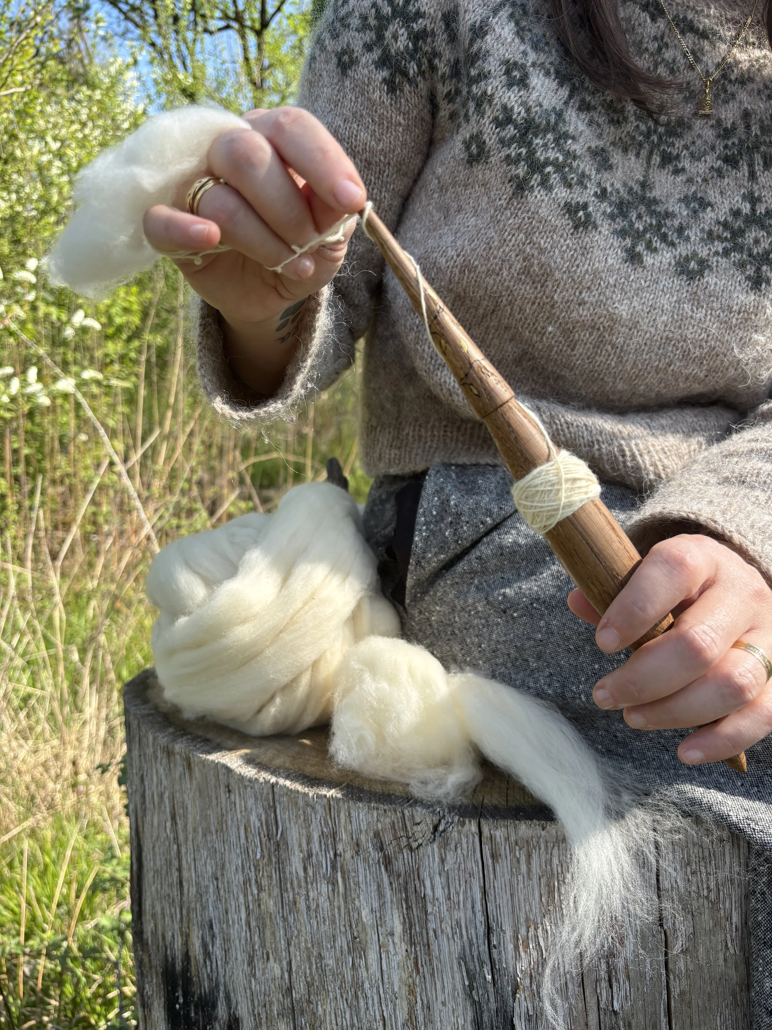 Una persona tiene un uncinetto lungo e di legno e sta lavorando a maglia con della lana bianca. Si trova all'aperto, vicino a un tronco d'albero, con foglie verdi e un cielo azzurro sullo sfondo.