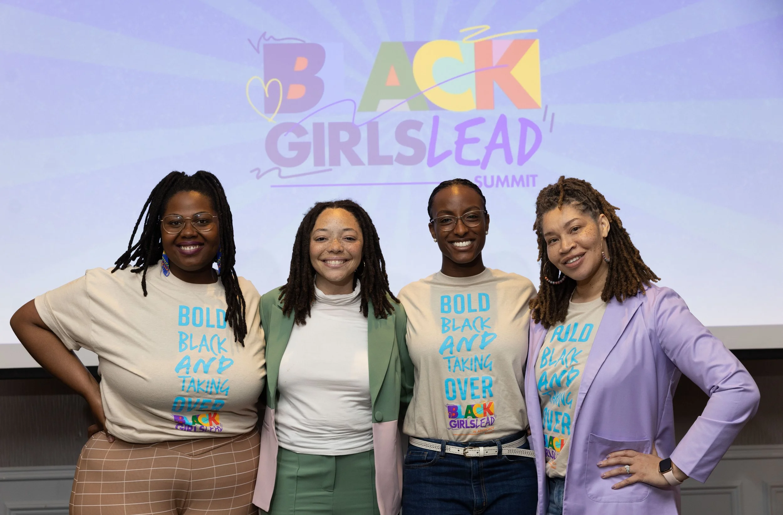 Four women standing together and smiling at a conference or event. They are wearing beige T-shirts with blue text and colorful accessories, standing in front of a screen with the text 'Back Girlslead Summit' in colorful letters.