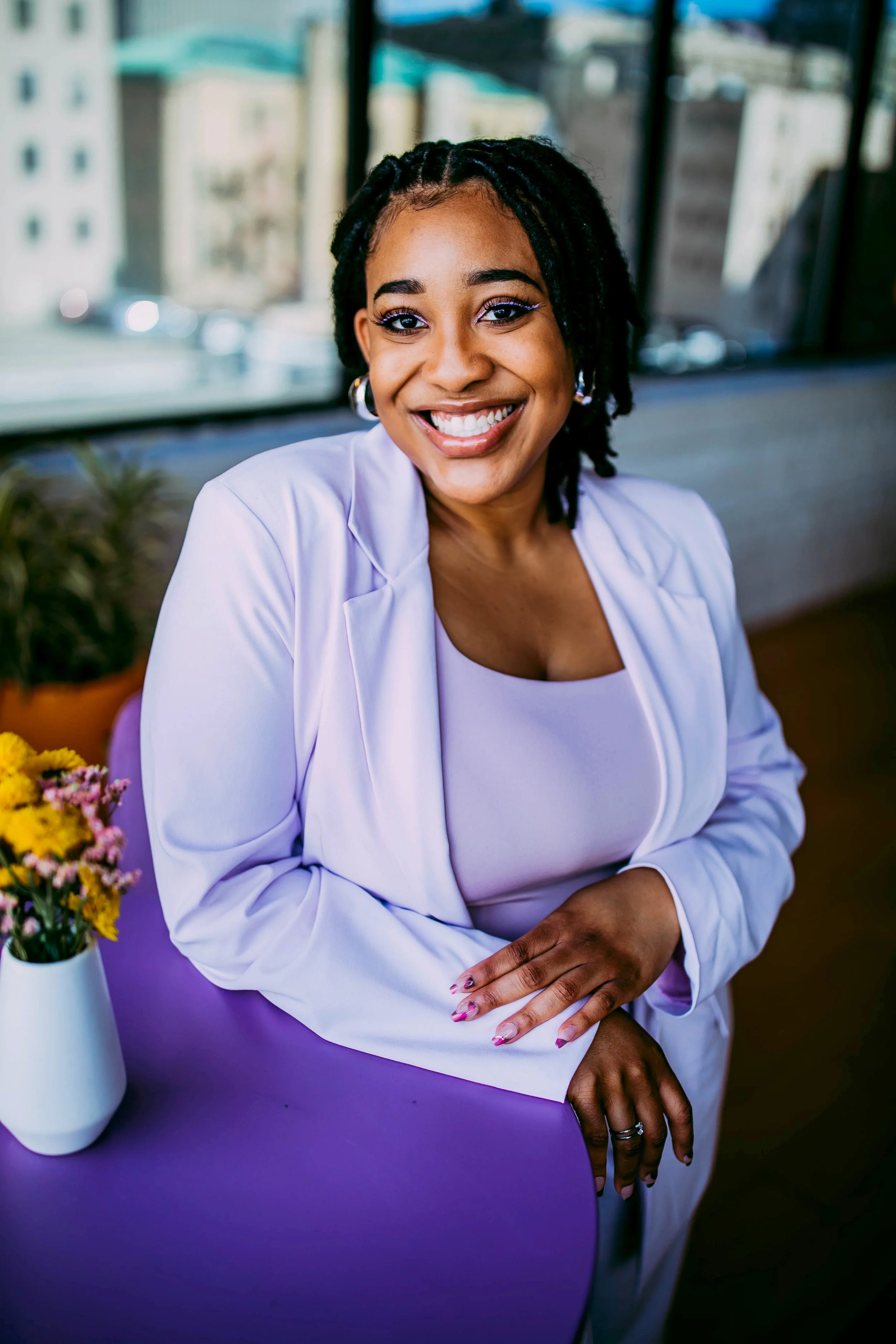 A smiling woman with dark skin and dreadlocks, wearing a white blazer, sitting at a purple table with a white vase of flowers, in front of a large window with city buildings in the background.