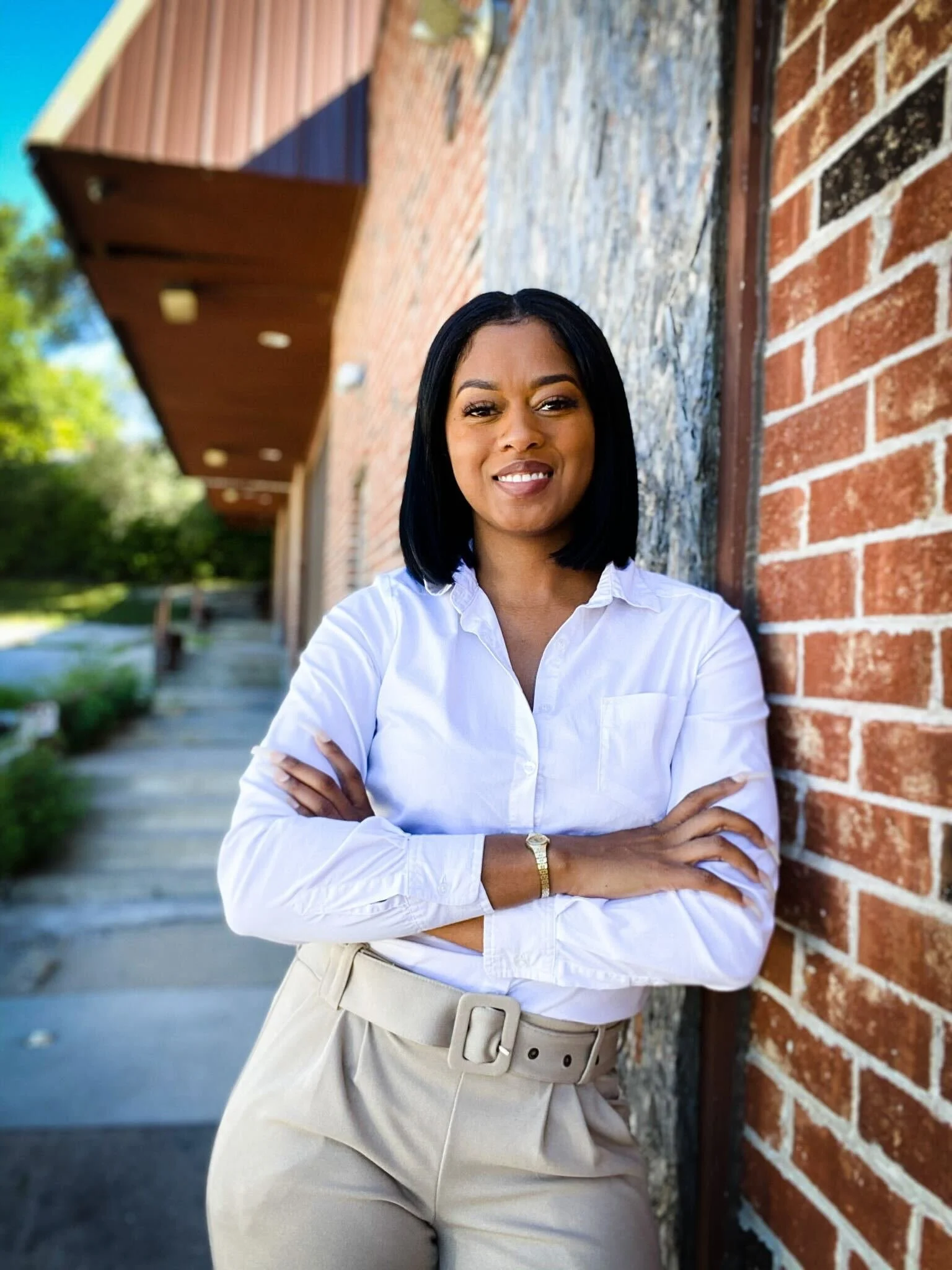 A woman with black hair, wearing a white shirt and beige pants, is standing outdoors against a red brick wall with her arms crossed and smiling.