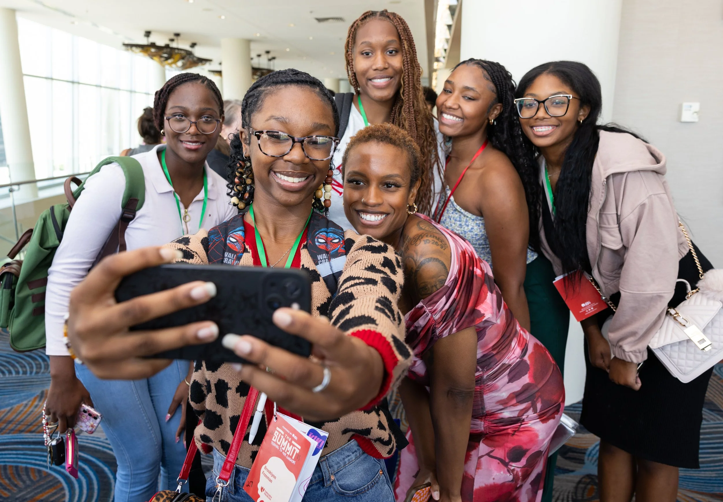 A group of seven women taking a selfie together at an indoor event, all smiling and looking at the phone camera.