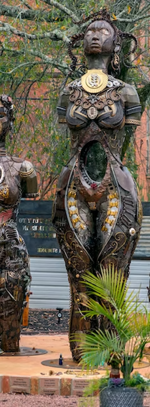 A tall wooden sculpture of a woman with intricate carvings and jewelry, surrounded by plants and other art pieces outdoors.