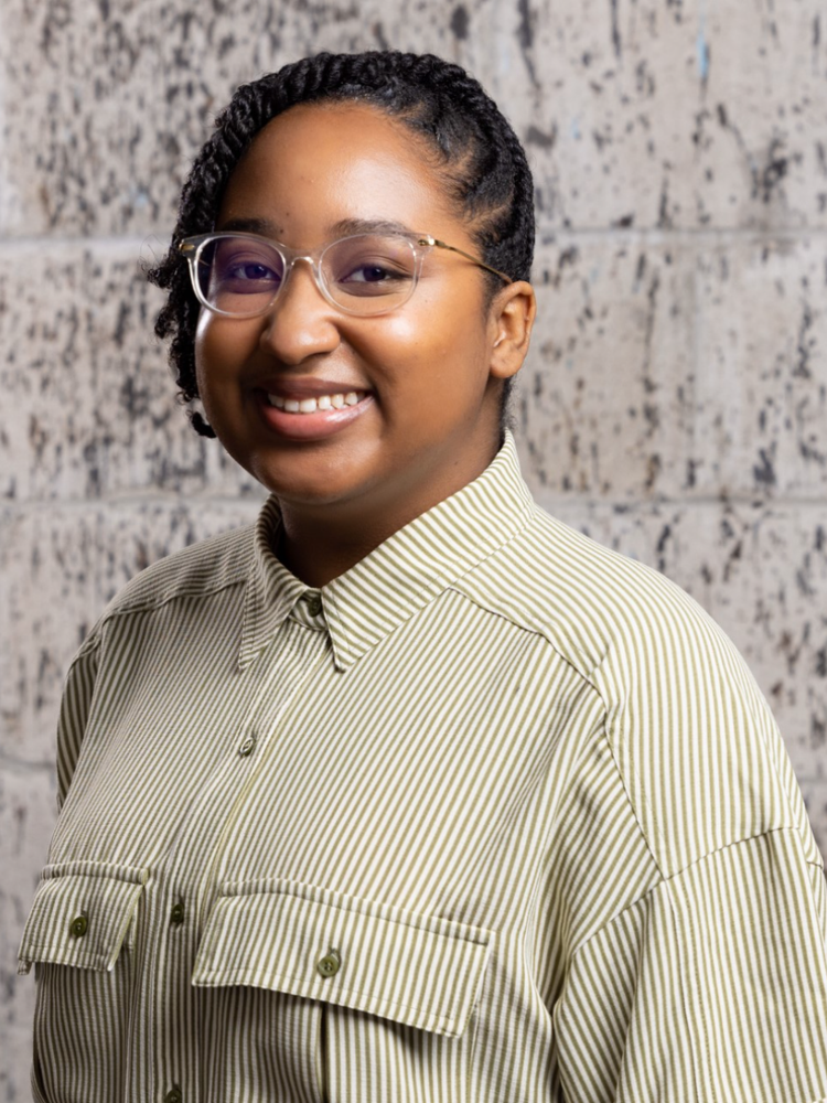 A woman with glasses smiling, standing in front of a brick wall, wearing a striped button-up shirt.