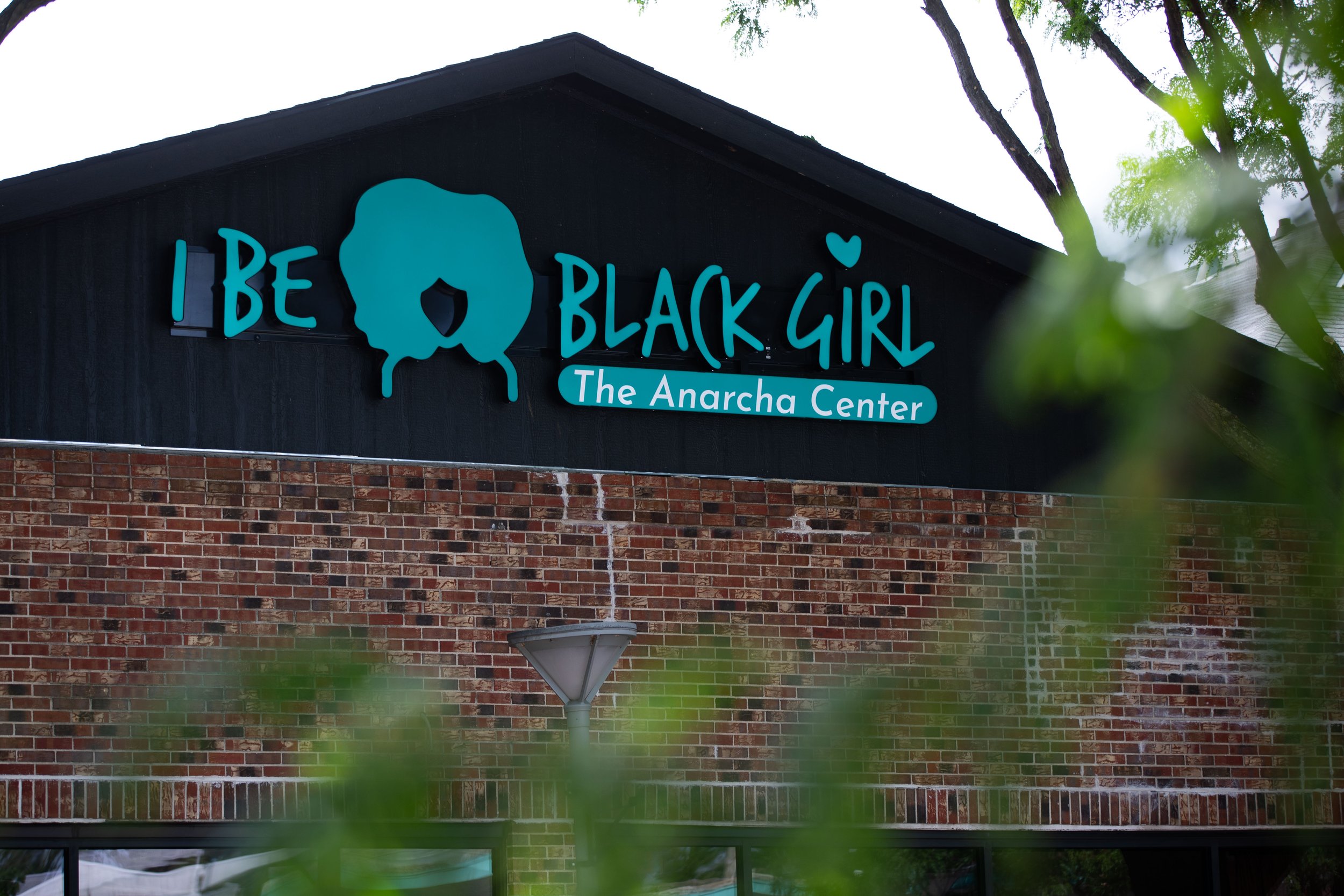 Anarcha Center in Omaha, Nebraska. I Be Black Girl' with a silhouette of a girl with curly hair, and smaller text underneath stating 'The Anarcha Center'. The building has black siding, a brick lower section, and trees partially visible around it.