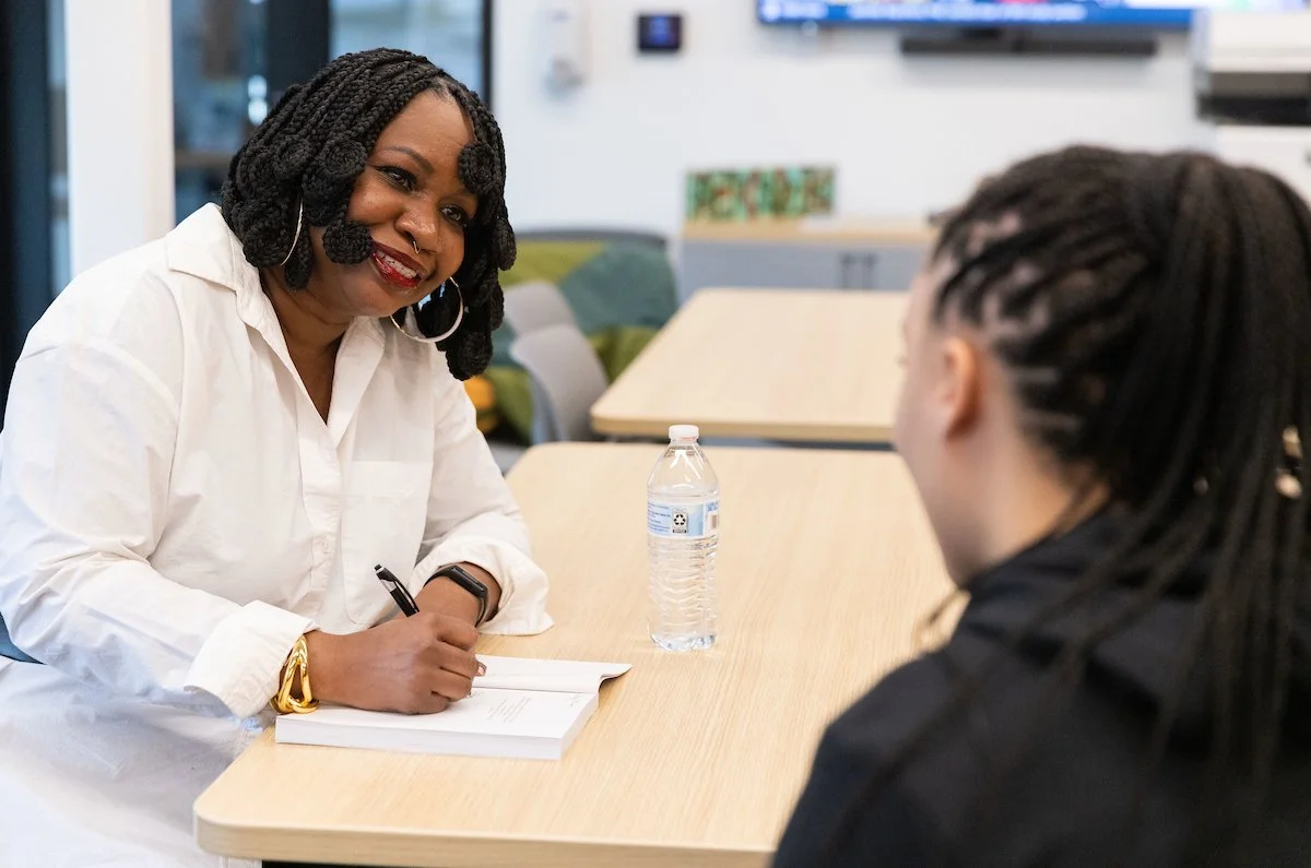 A woman with styled dreadlocks and hoop earrings smiling during a conversation across a table in a bright room, with a water bottle on the table.