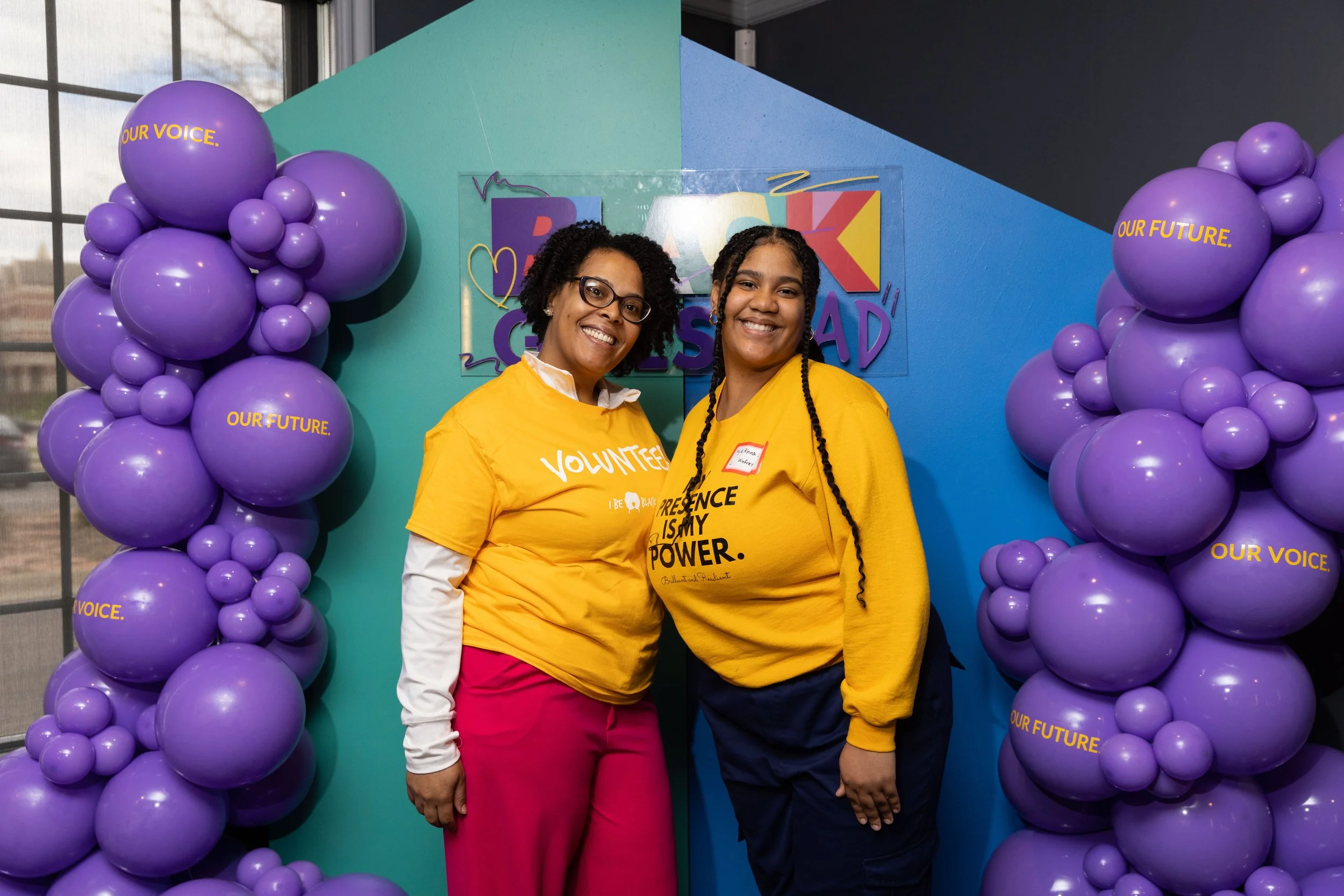 Two women smiling and posing at a colorful event decorated with purple balloons that have the words "OUR VOICE" and "OUR FUTURE" written on them. The woman on the left has curly hair, glasses, and is wearing a yellow shirt with white and pink pants. The woman on the right has braided hair and is wearing a yellow shirt with black pants.